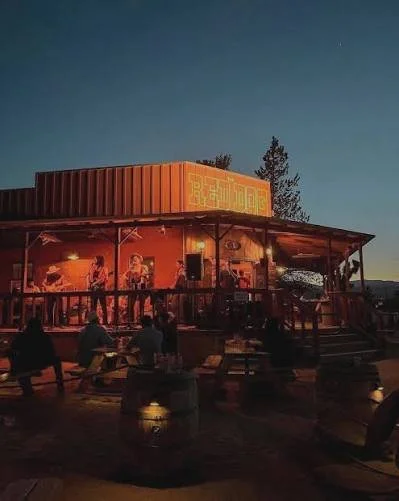 Outdoor evening scene of a music venue made from shipping containers with people sitting at tables and a band performing on stage.