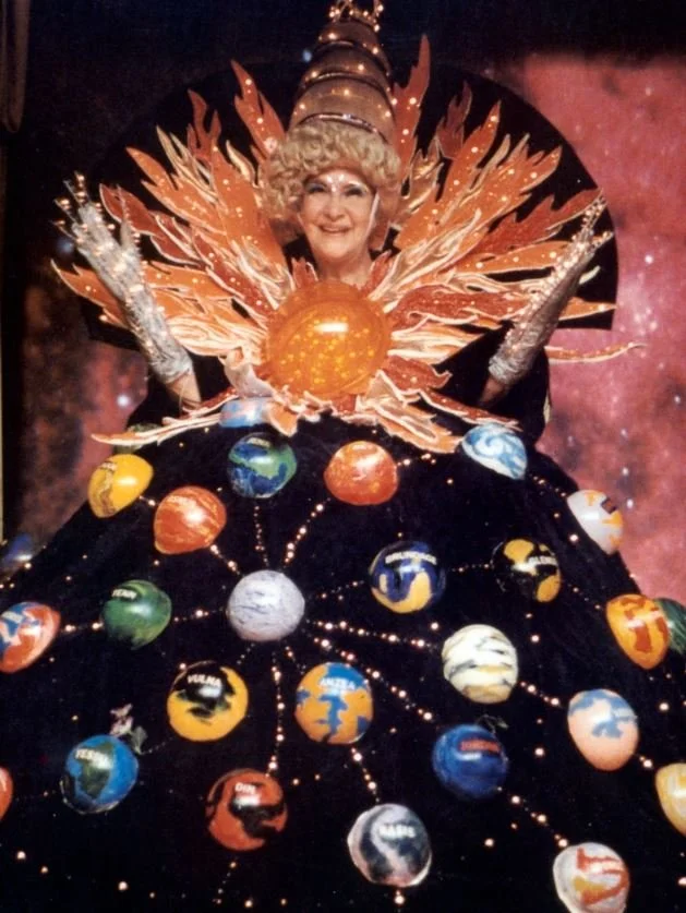 Ruth E. Norman Uriel wearing an elaborate space-themed costume with planets and space elements, posing in front of a starry background.