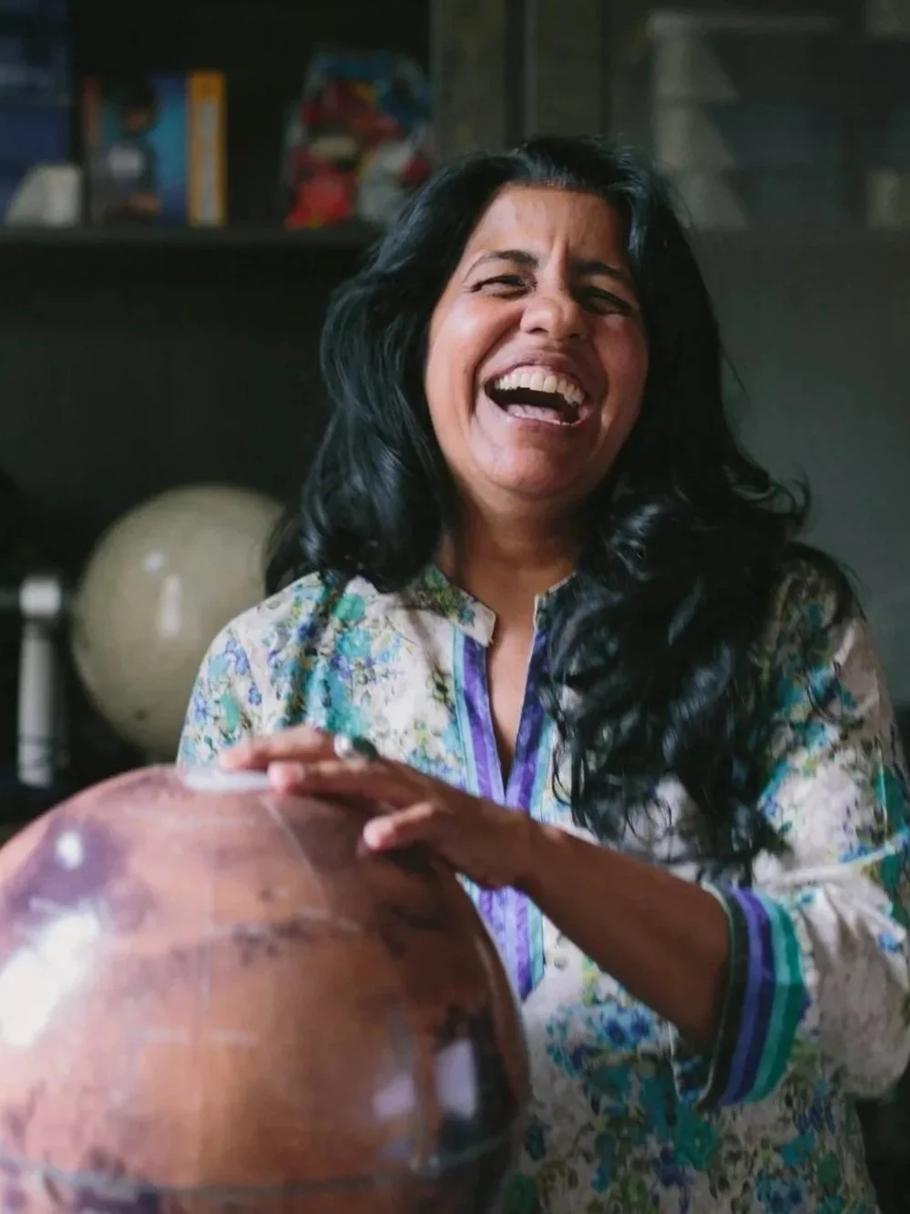 Dr. Aparna Venkatesan long dark hair laughing, wearing a colorful floral shirt, in a room with shelves and pottery in the background.