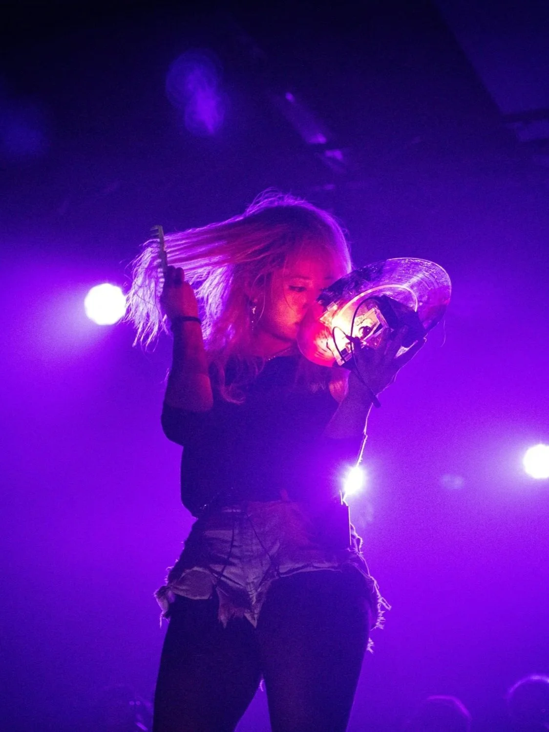 Performer with blonde hair holding a lighted device on stage illuminated by purple and white lights.