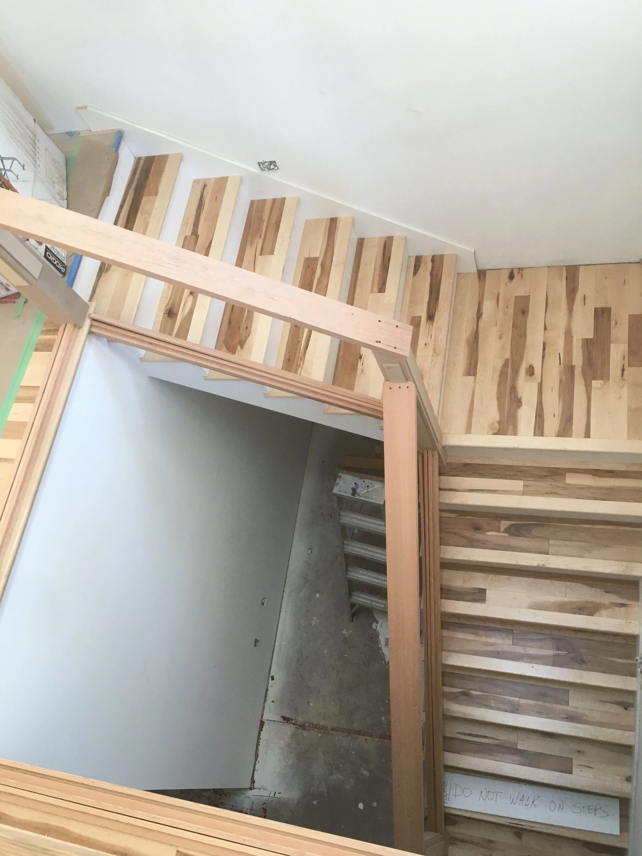 Interior view of a staircase under construction with wooden steps, a protective white wall on the left, and a partially finished wall on the right with horizontal wooden panels. A handwritten sign on the bottom step reads, 'DO NOT WALK ON STEPS'.