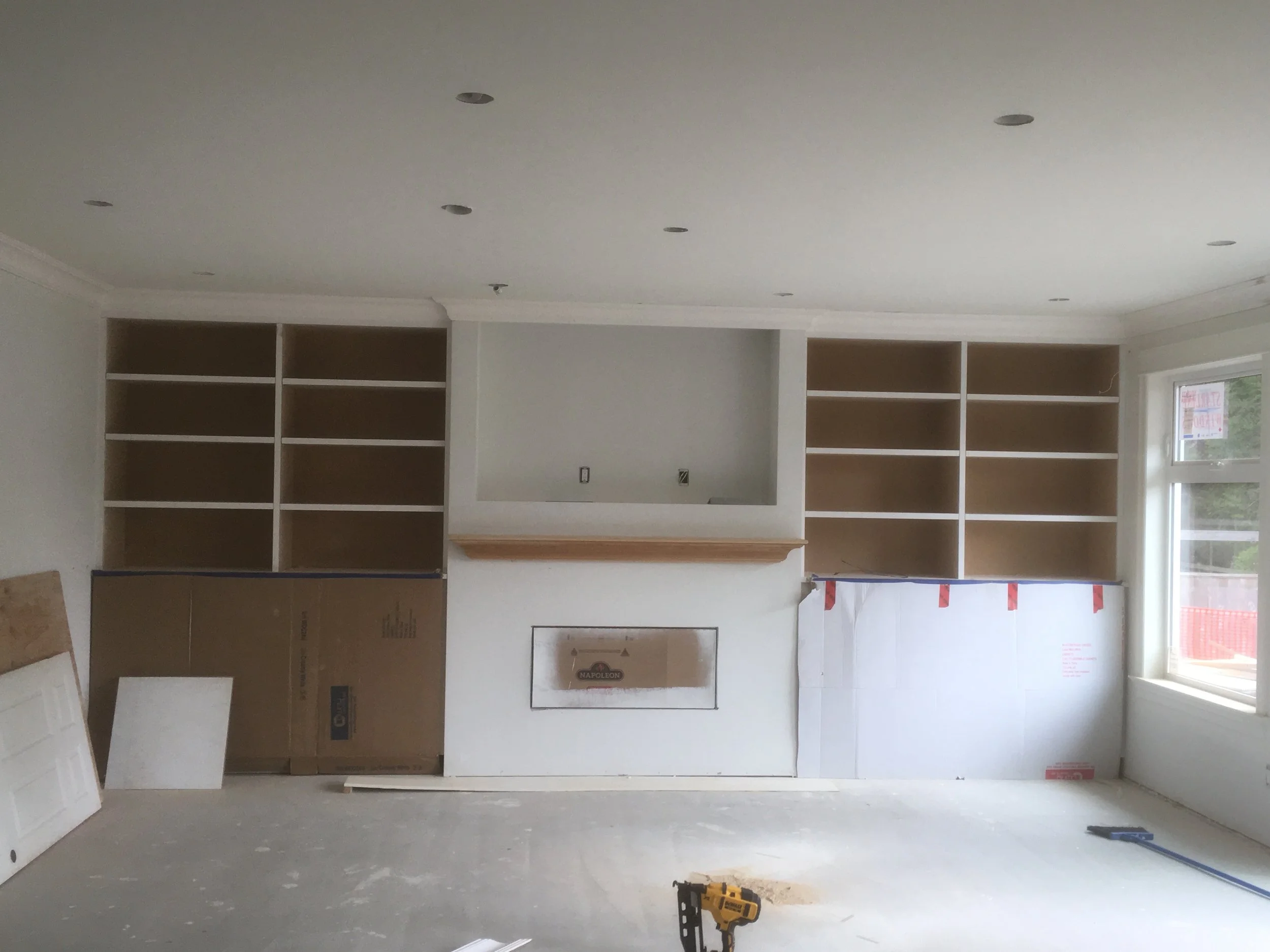 Interior of a room under construction with built-in bookshelves, a fireplace, and two large windows. Tools are on the floor.