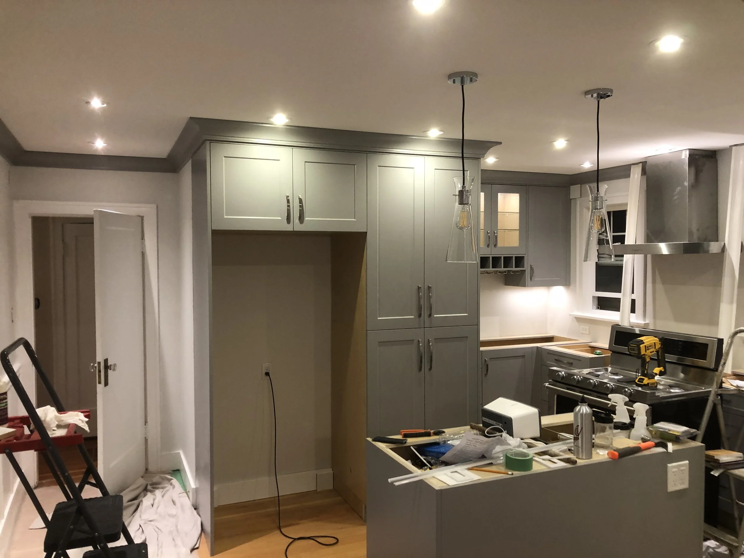 A kitchen under renovation with light green cabinets, a stove, and construction tools and supplies on the countertop. Two pendant lights hang from the ceiling.
