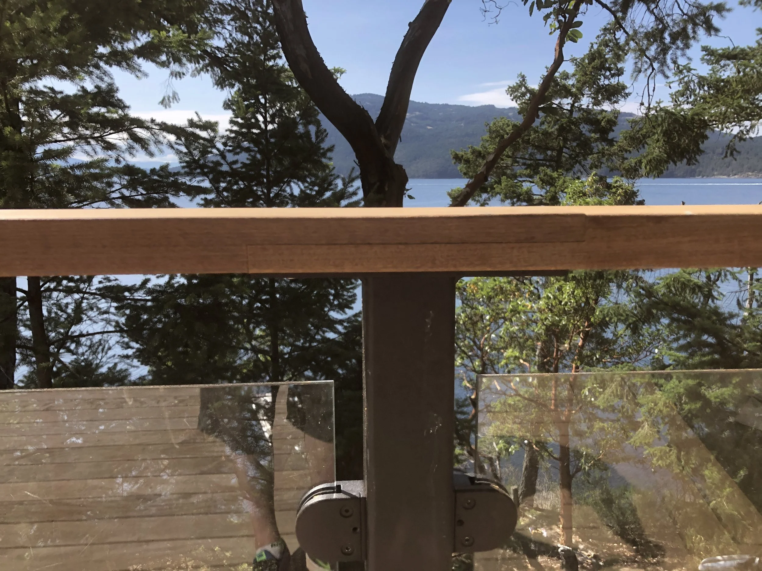 Photo of a waterfront view with trees, mountains, and a blue sky, taken through a window with a wooden railing and a metal latch.
