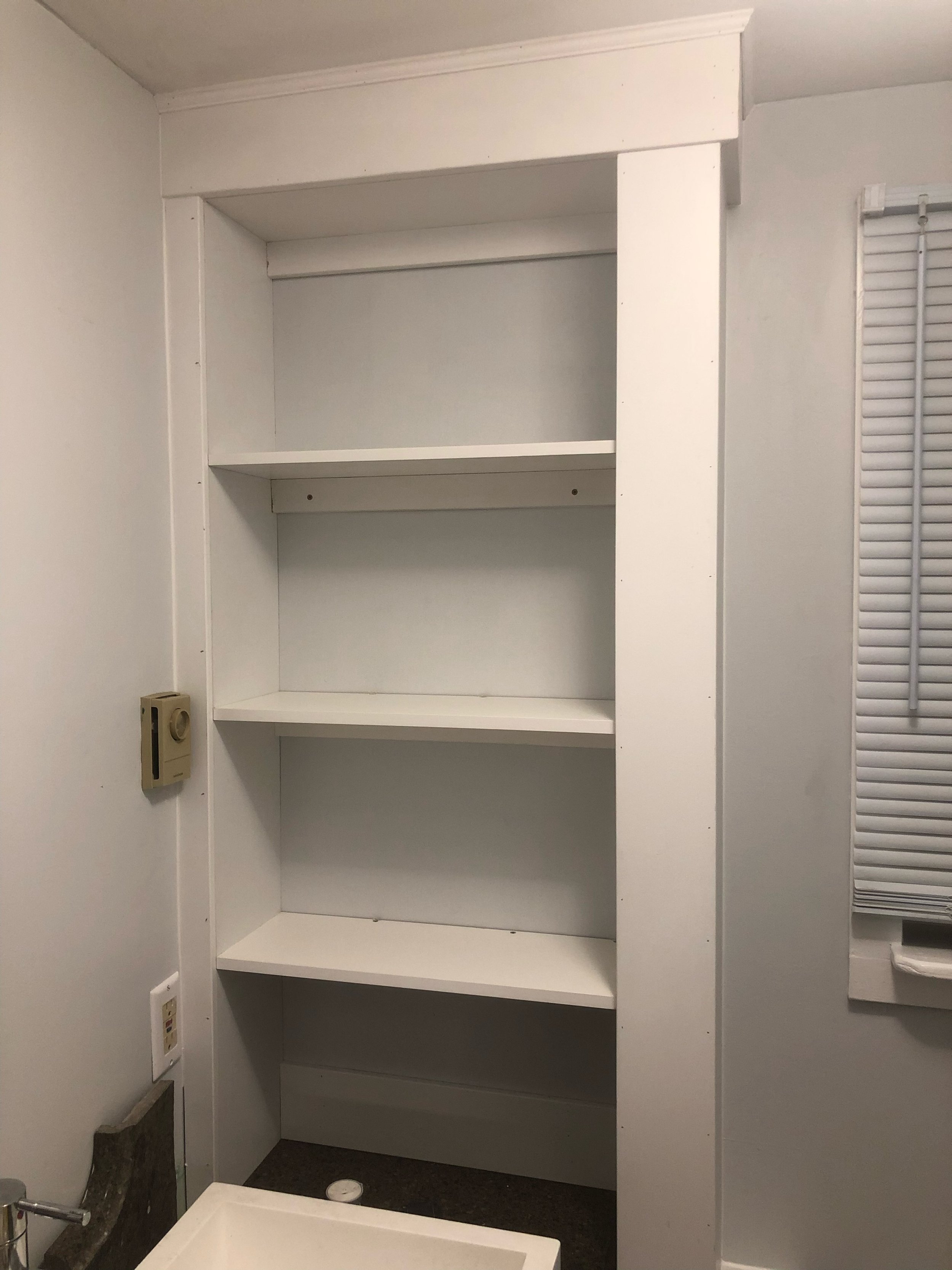 Empty white shelving unit with four shelves in a room with white walls and a dark floor, near a small sink and a window with closed blinds.