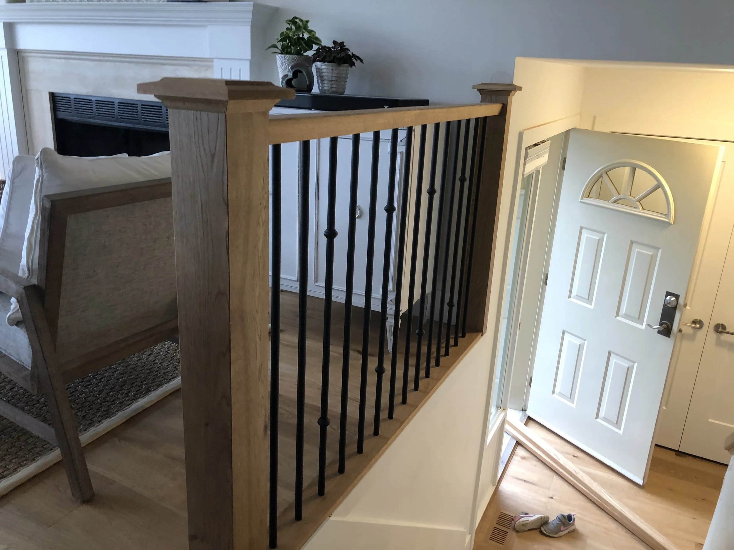 View of a staircase with wooden handrail and black metal balusters leading to a doorway with a fan and a wall with a potted plant on top.