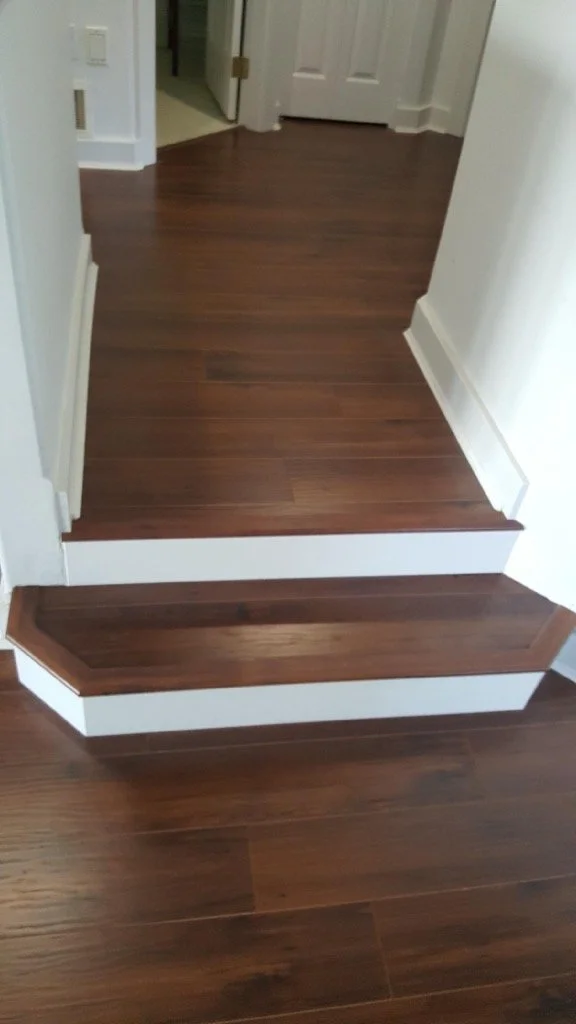 A small set of two wooden steps with white trim in a hallway with hardwood flooring and a white door at the end.