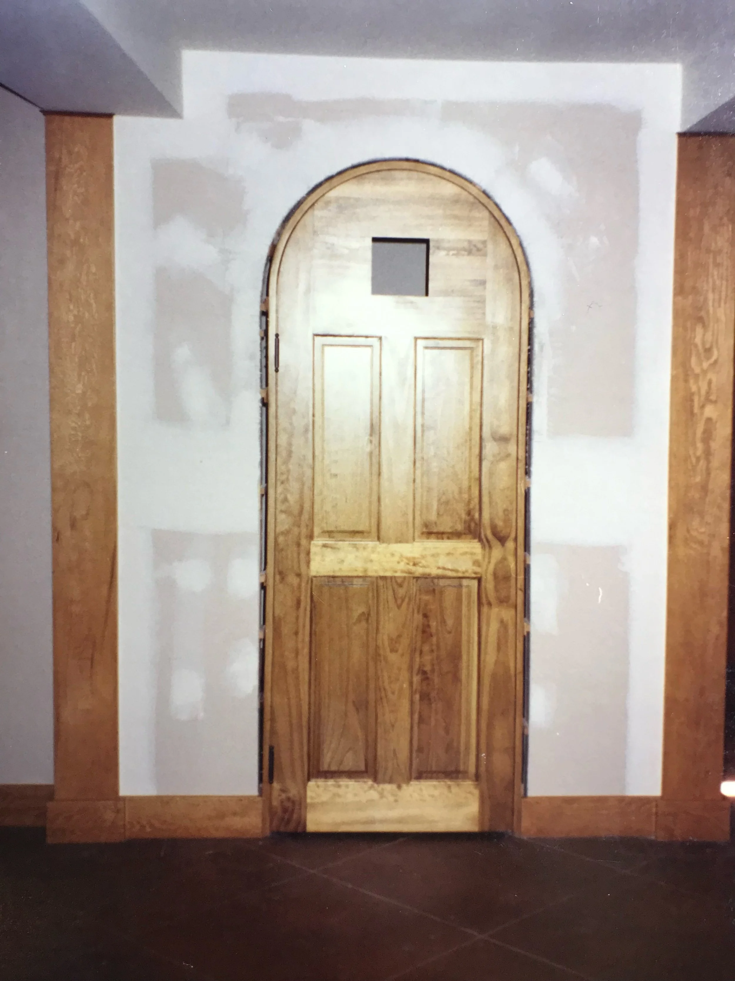 Wooden door with a small window at the top, installed within a wall under construction or renovation, with unfinished drywall surrounding it.