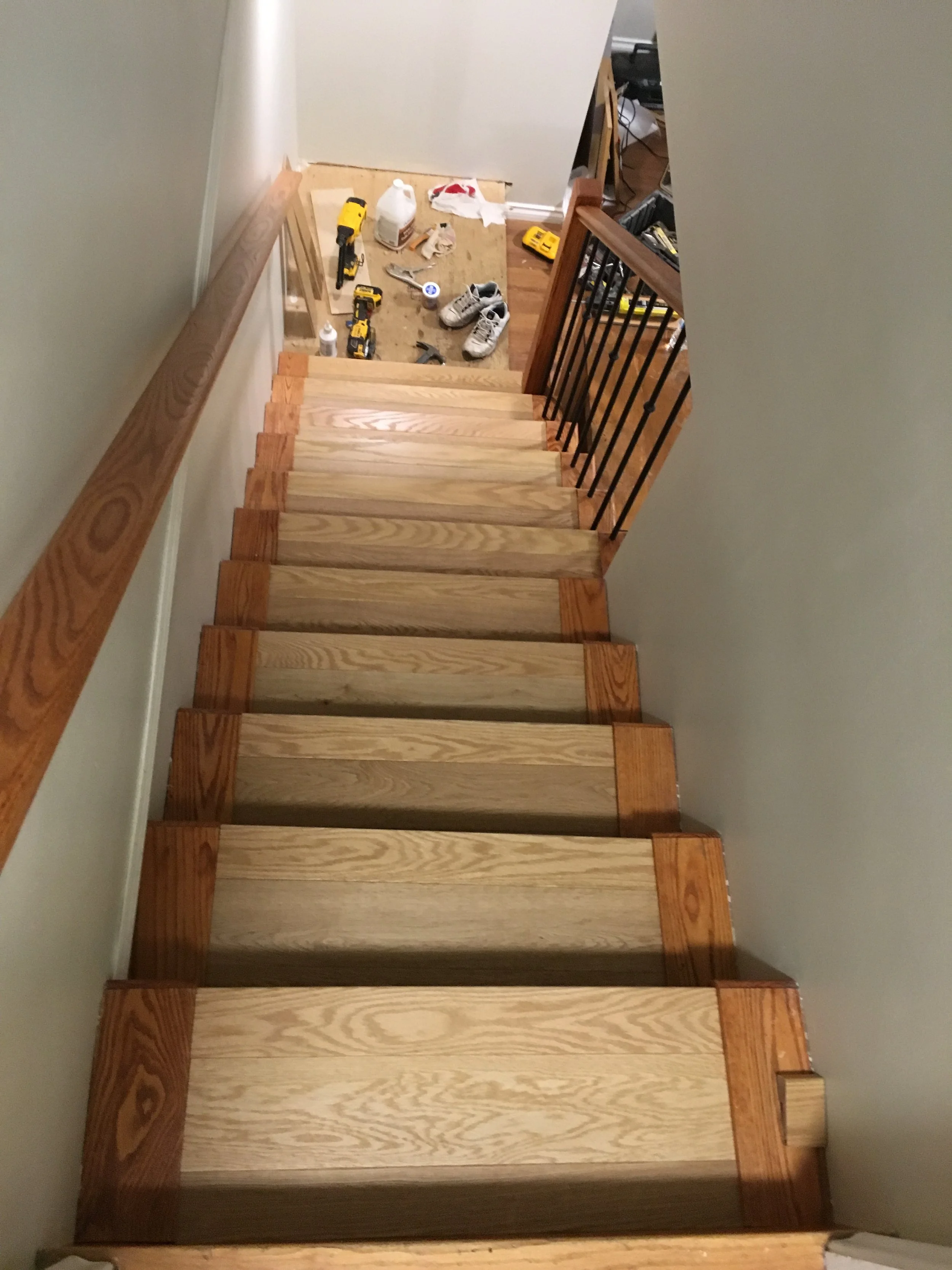View from the top of a staircase looking down, showing wooden stairs, black metal railing, and a small landing area with tools and shoes, indicating ongoing staircase construction or renovation.