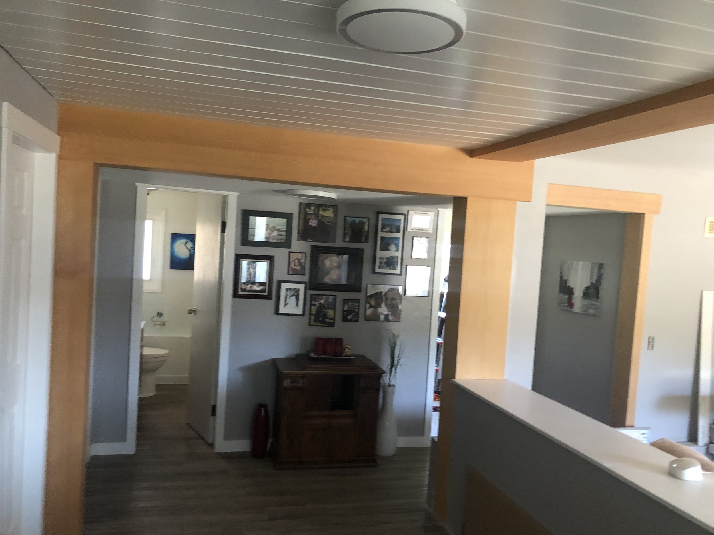 Interior view of a house with a hallway leading to a room that has framed pictures on the wall, a wooden cabinet, and a window with sunlight.