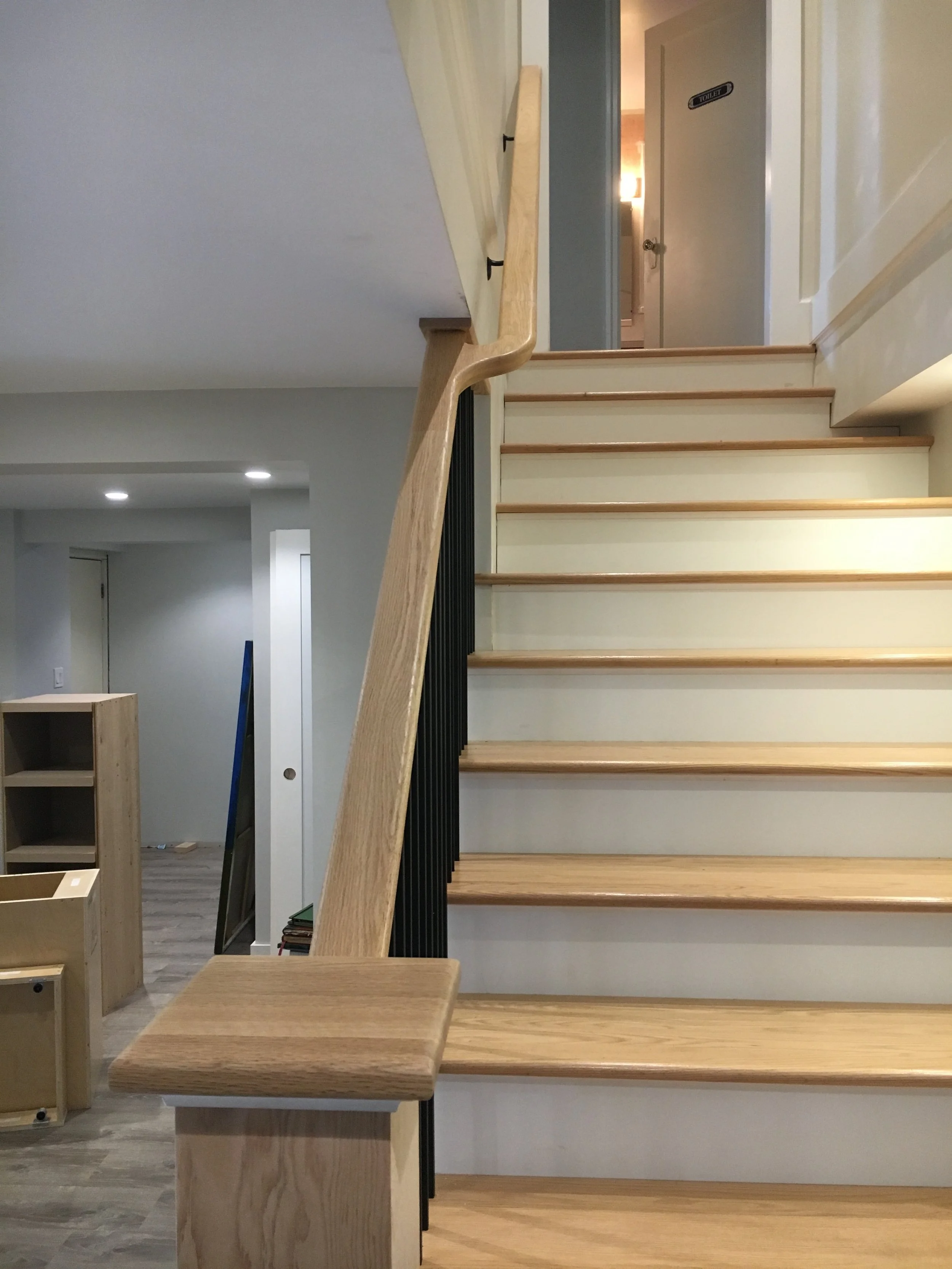 Interior view of a staircase with wooden steps and a light-colored handrail, leading to a hallway with a door labeled 'Toilet' and visible room furnishings.
