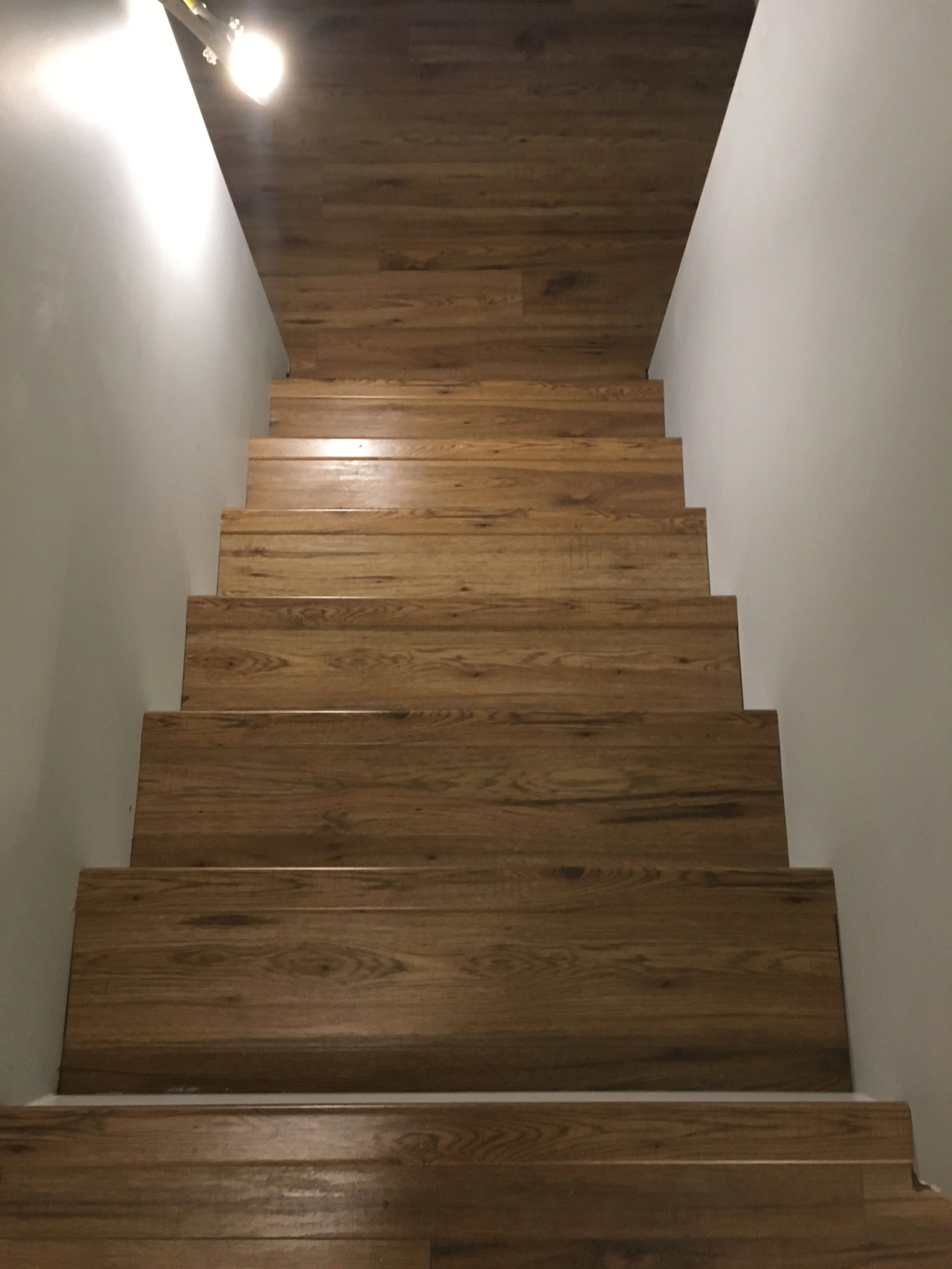 Wooden staircase with a white wall on one side and a gray wall on the other, illuminated by a ceiling light.