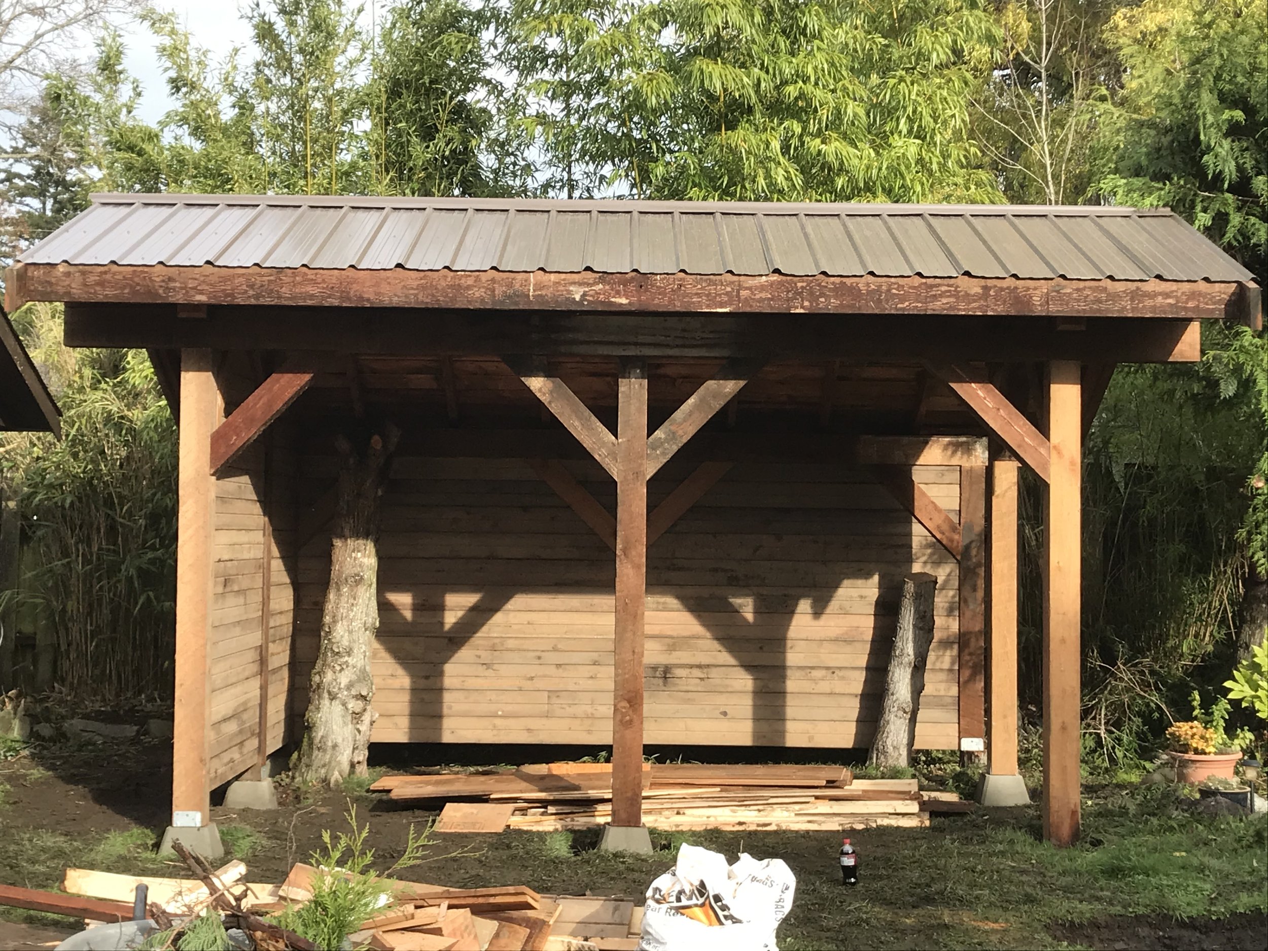 A wooden structure under construction with vertical support beams and a metal roof, surrounded by trees and construction materials on the ground.
