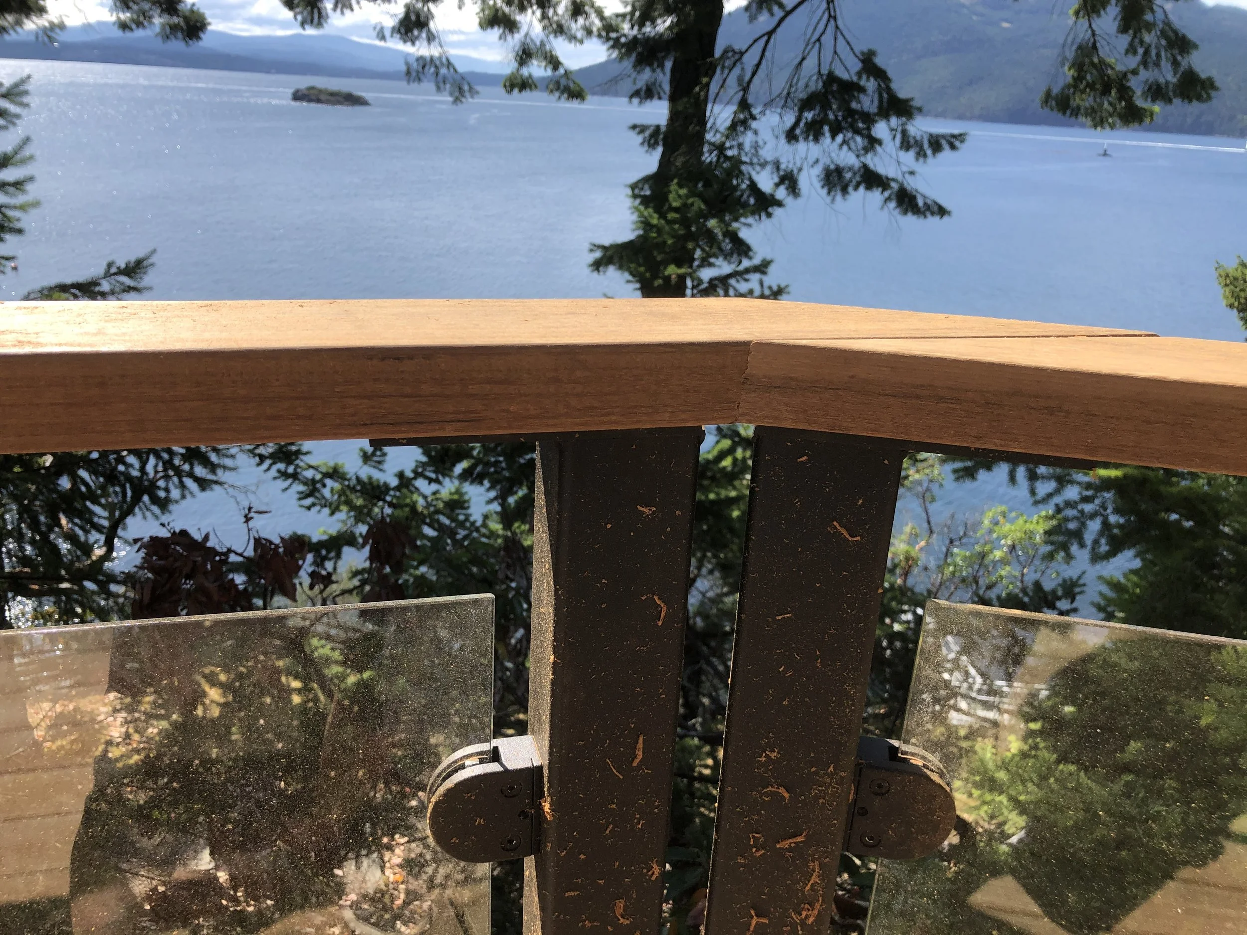 View of a large lake or river with small island, surrounded by mountains, seen through a wooden railing and trees.