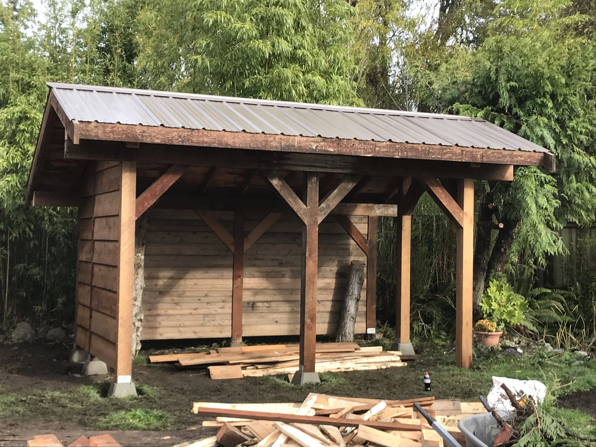 A wooden shelter under construction outdoors, surrounded by green trees and plants, with building materials and tools on the ground.
