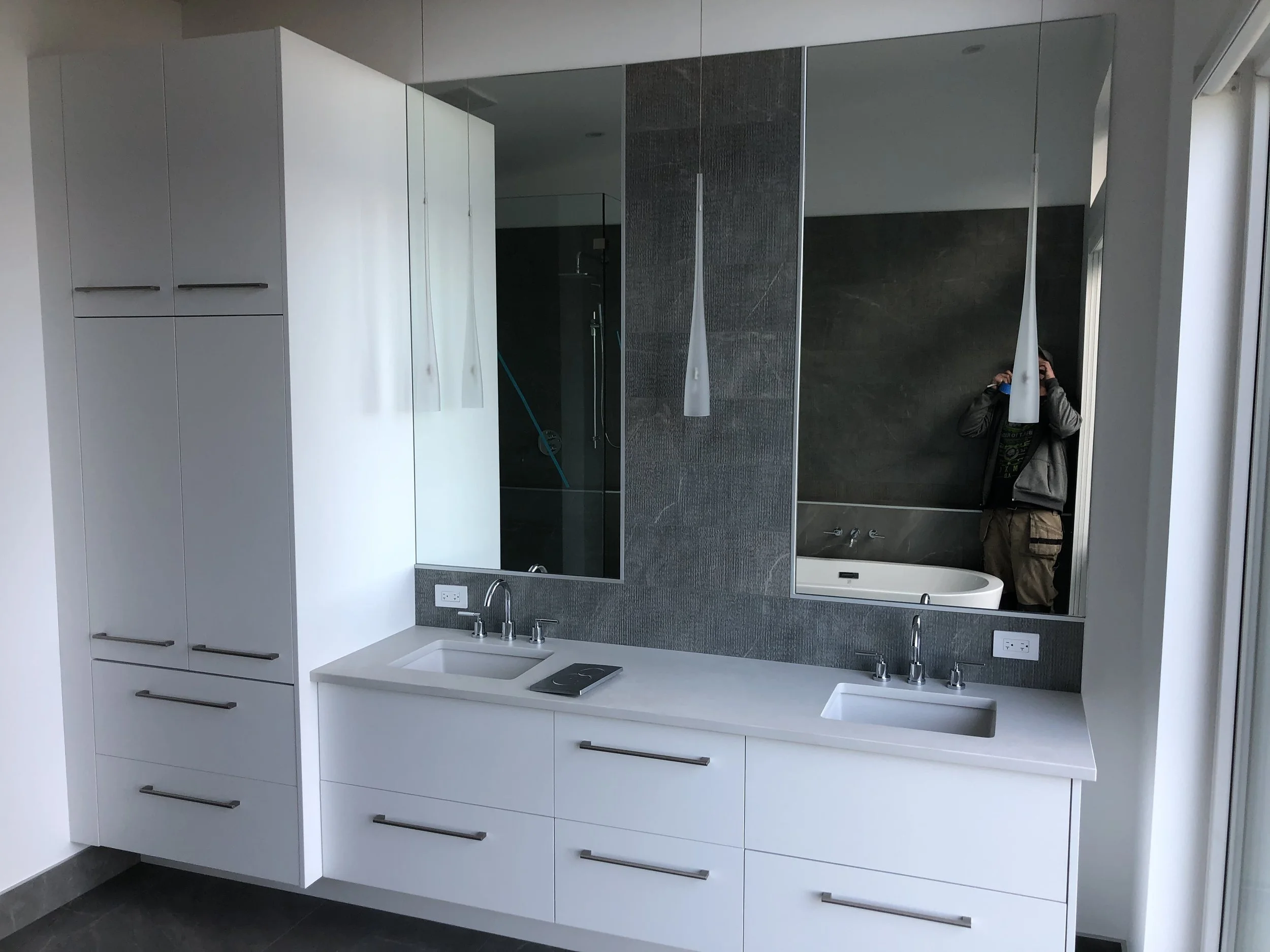 Modern bathroom with white cabinetry, a large mirror, a small sink, and a shower area with a glass door, a person taking a photo in the mirror, and a gray tiled wall.