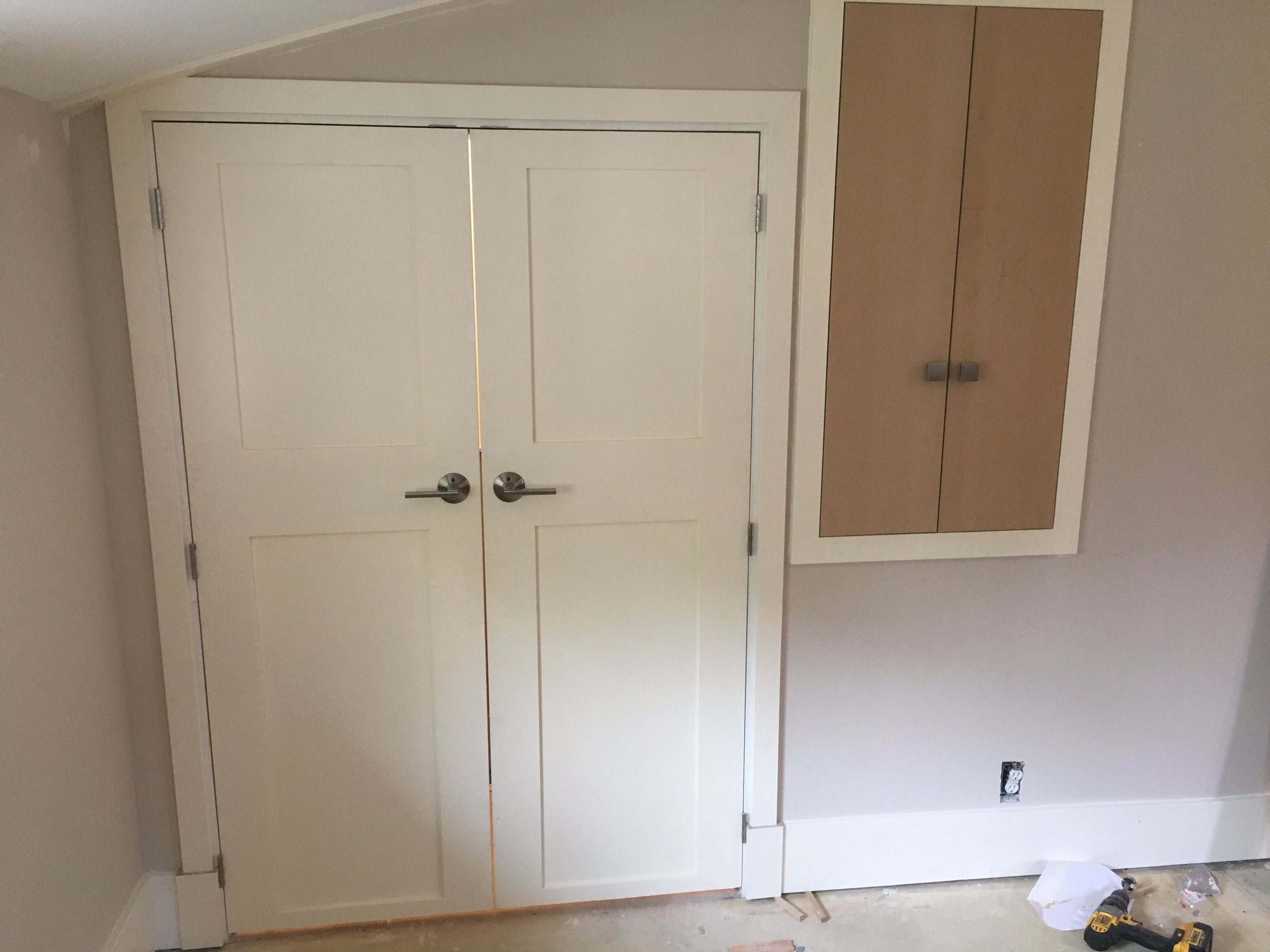 Interior view of a room under construction or renovation, showing installed white double doors with metallic handles, a wooden cabinet to the right, and an unfinished electrical outlet near the floor with tools and construction materials below.