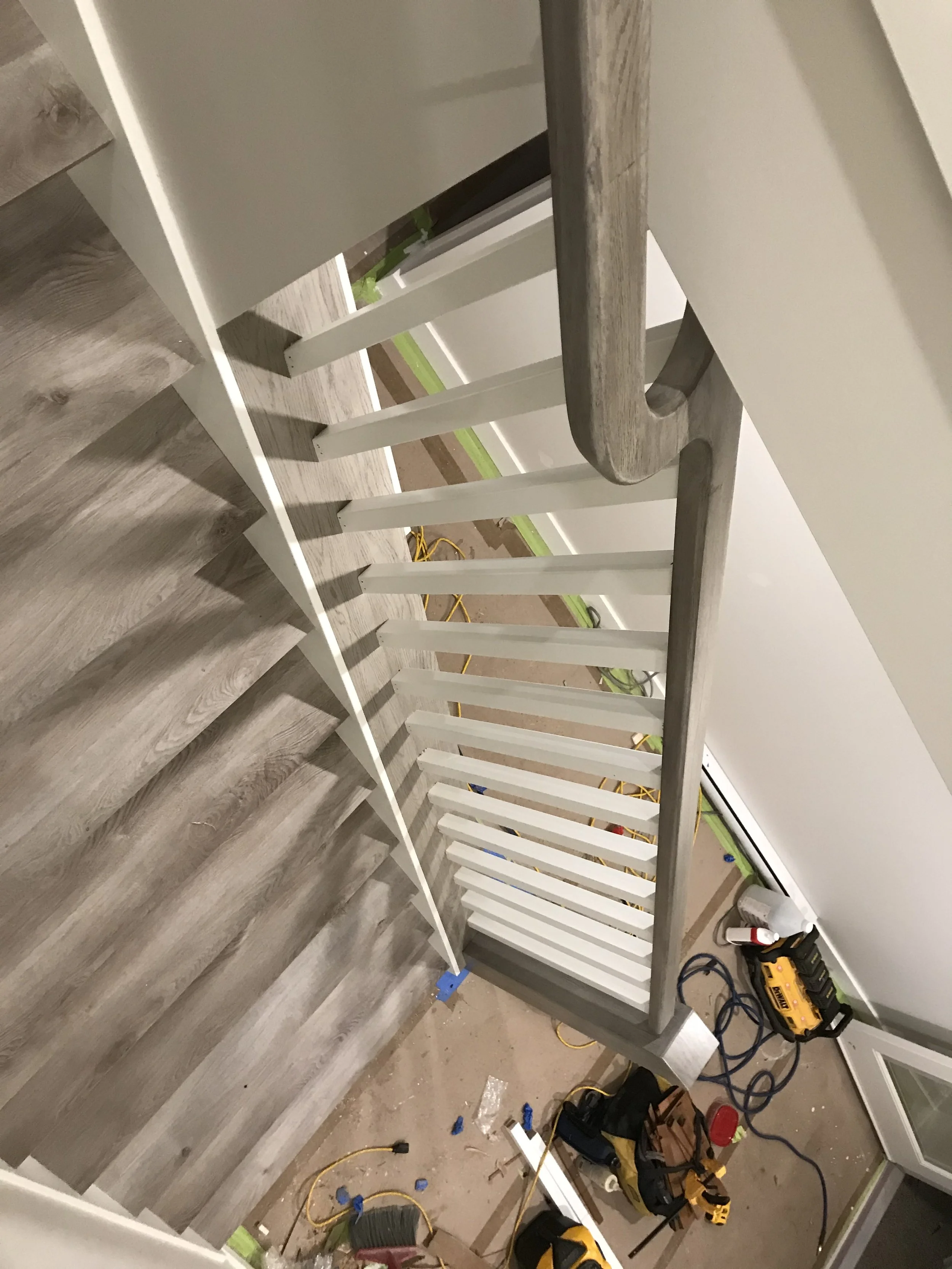 The image shows a partially constructed or assembled staircase railing with a wooden handrail and white vertical balusters. The surrounding area has construction tools, materials, and loose wires on the floor.