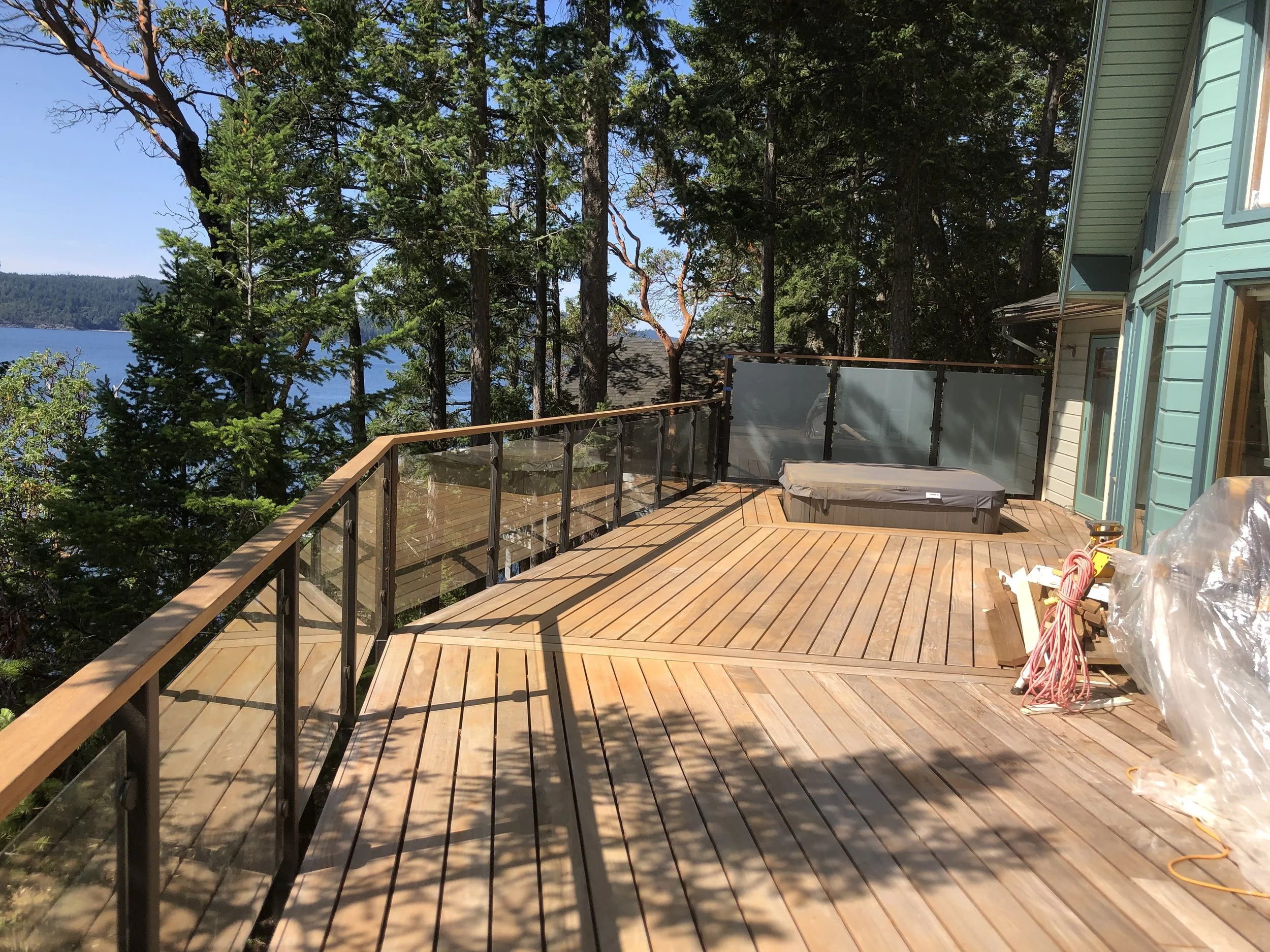 A wooden deck with a hot tub, surrounded by a railing, overlooking trees and a body of water, with a house exterior and construction materials visible.