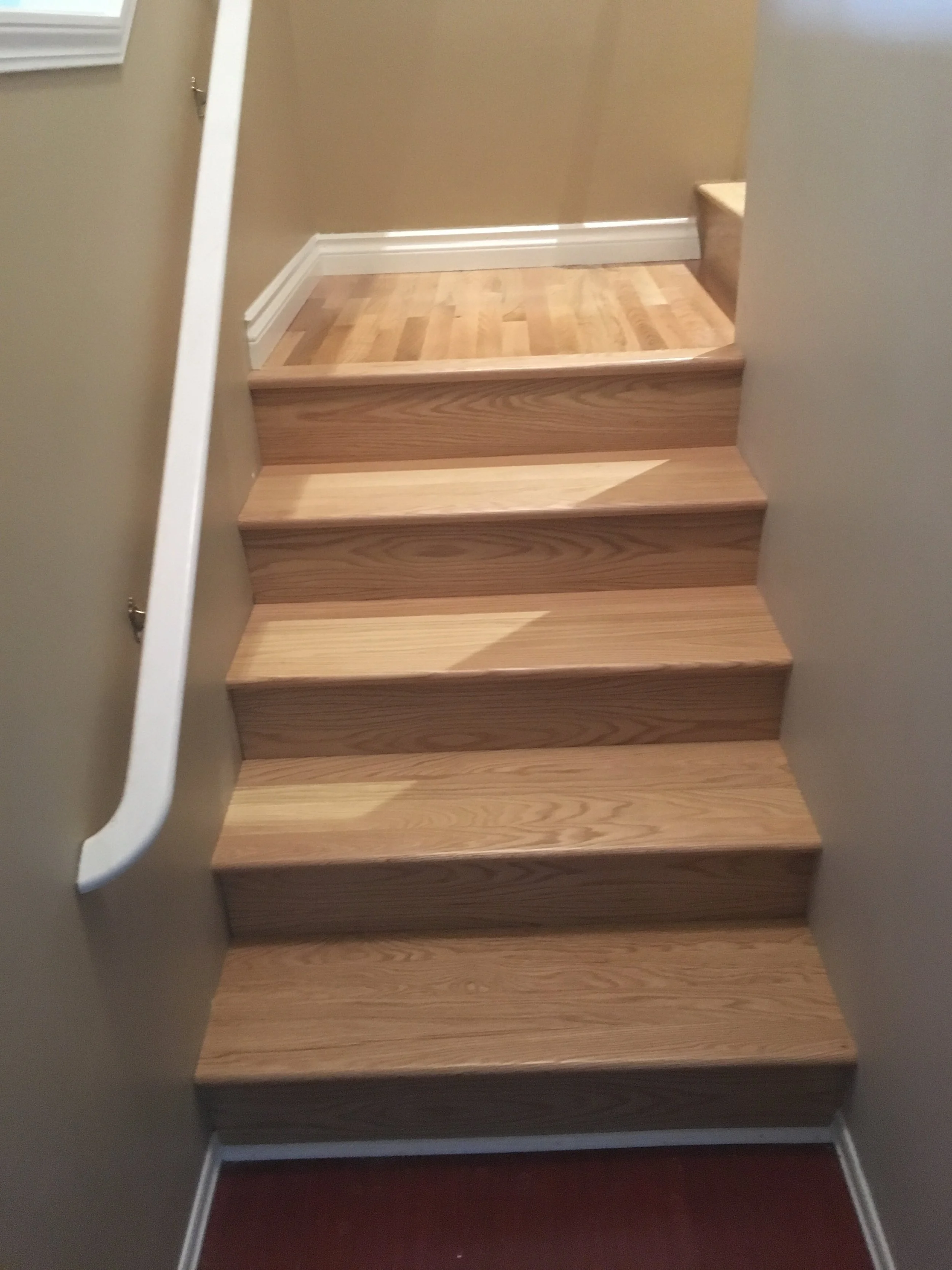 A staircase with six wooden steps in a home interior. The steps are stained a light natural wood, with white trim along the edges. To the right, there is a small landing with a carpeted surface, and a partial view of a wall. A white handrail is visib