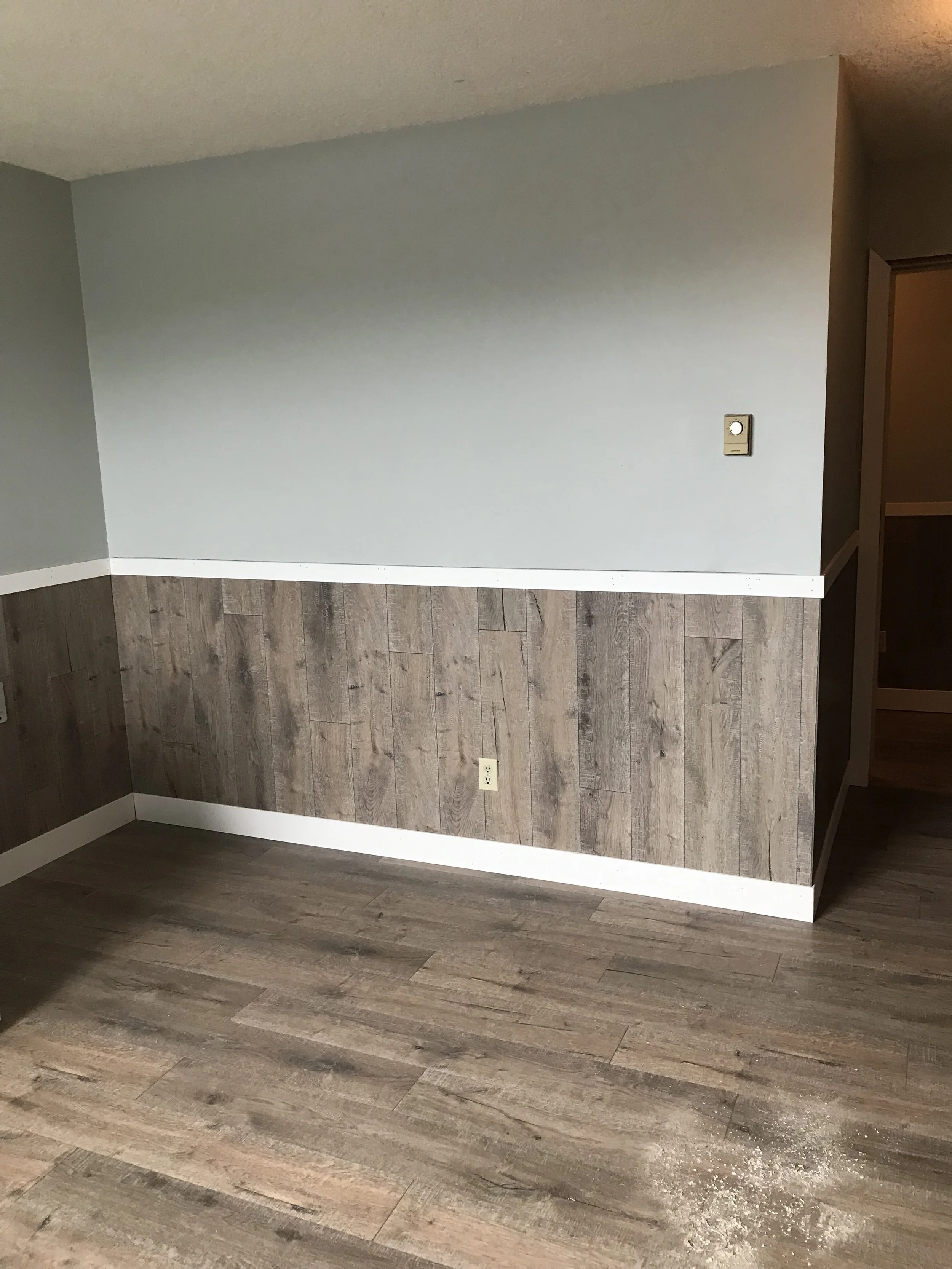 Interior corner of a room with a light gray wall, wood paneling halfway up, and wood flooring; electrical outlet near the floor and a thermostat on the wall.