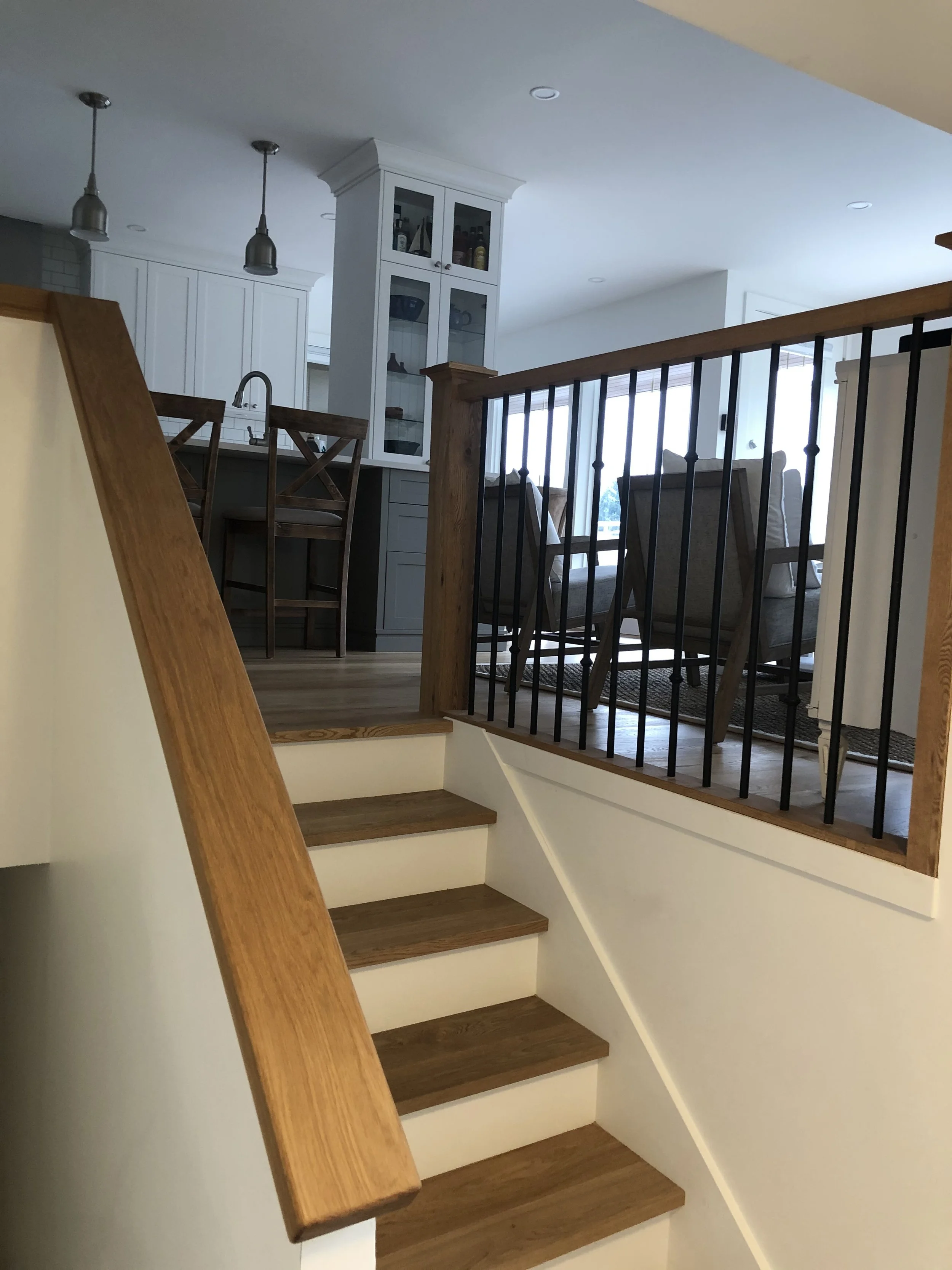Image of a staircase with wooden steps and a wooden handrail, leading to an open area with a white kitchen and living space, including chairs and a cabinet.