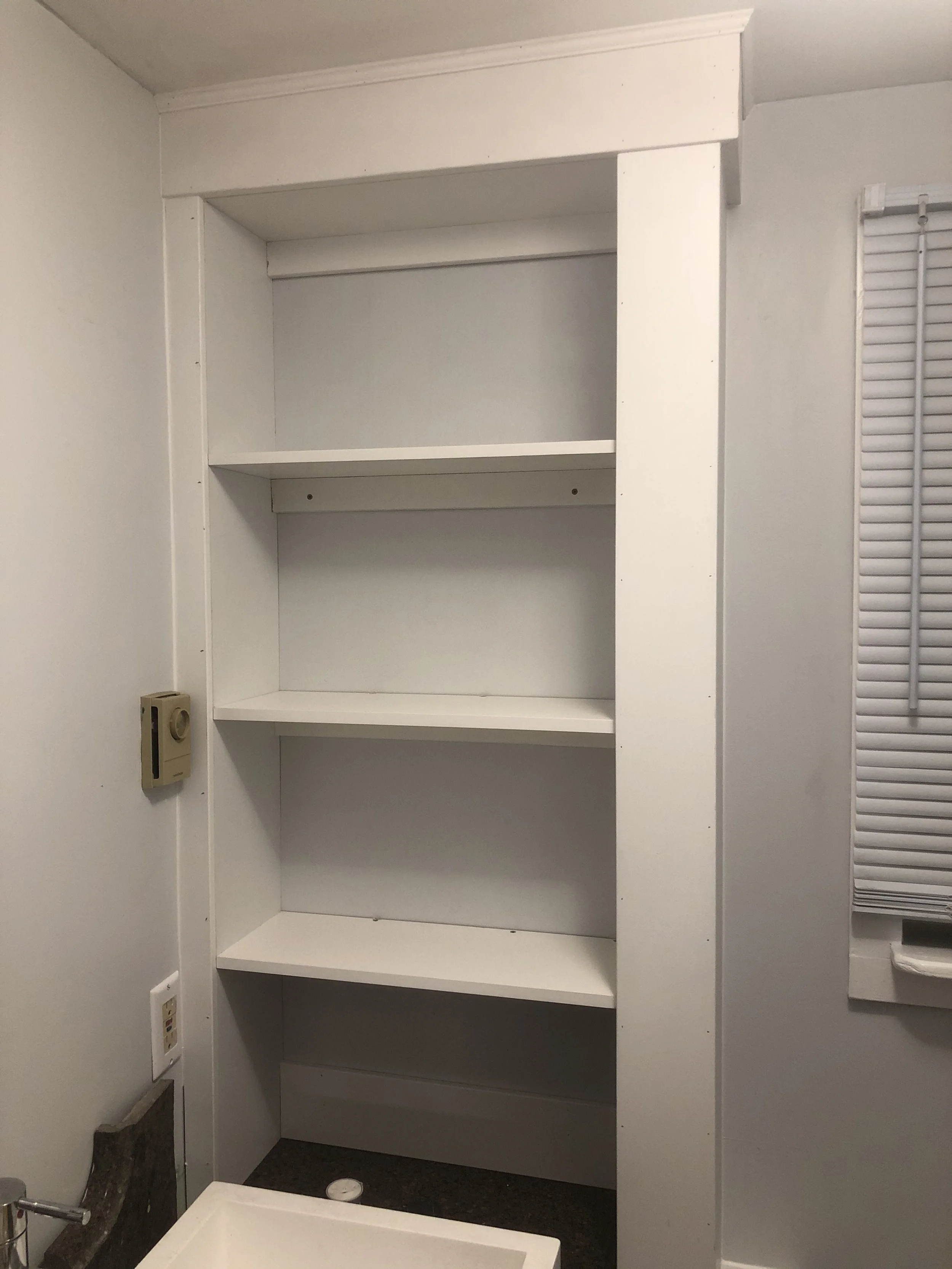 Empty white built-in shelving unit with four shelves in a room, next to a window with white horizontal blinds.