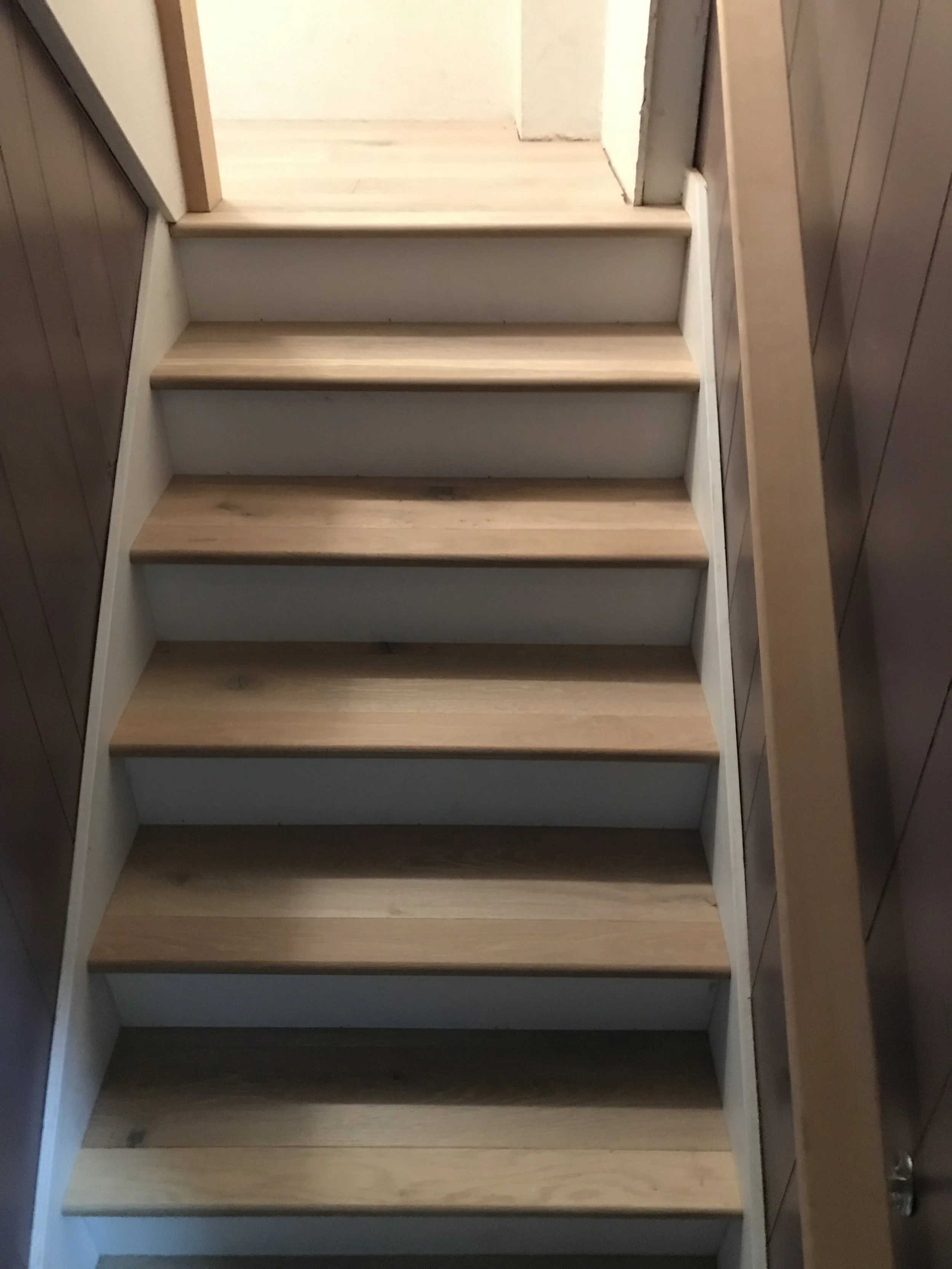 A narrow staircase with wooden steps, white side supports, and dark wood paneling on the walls, leading upwards.