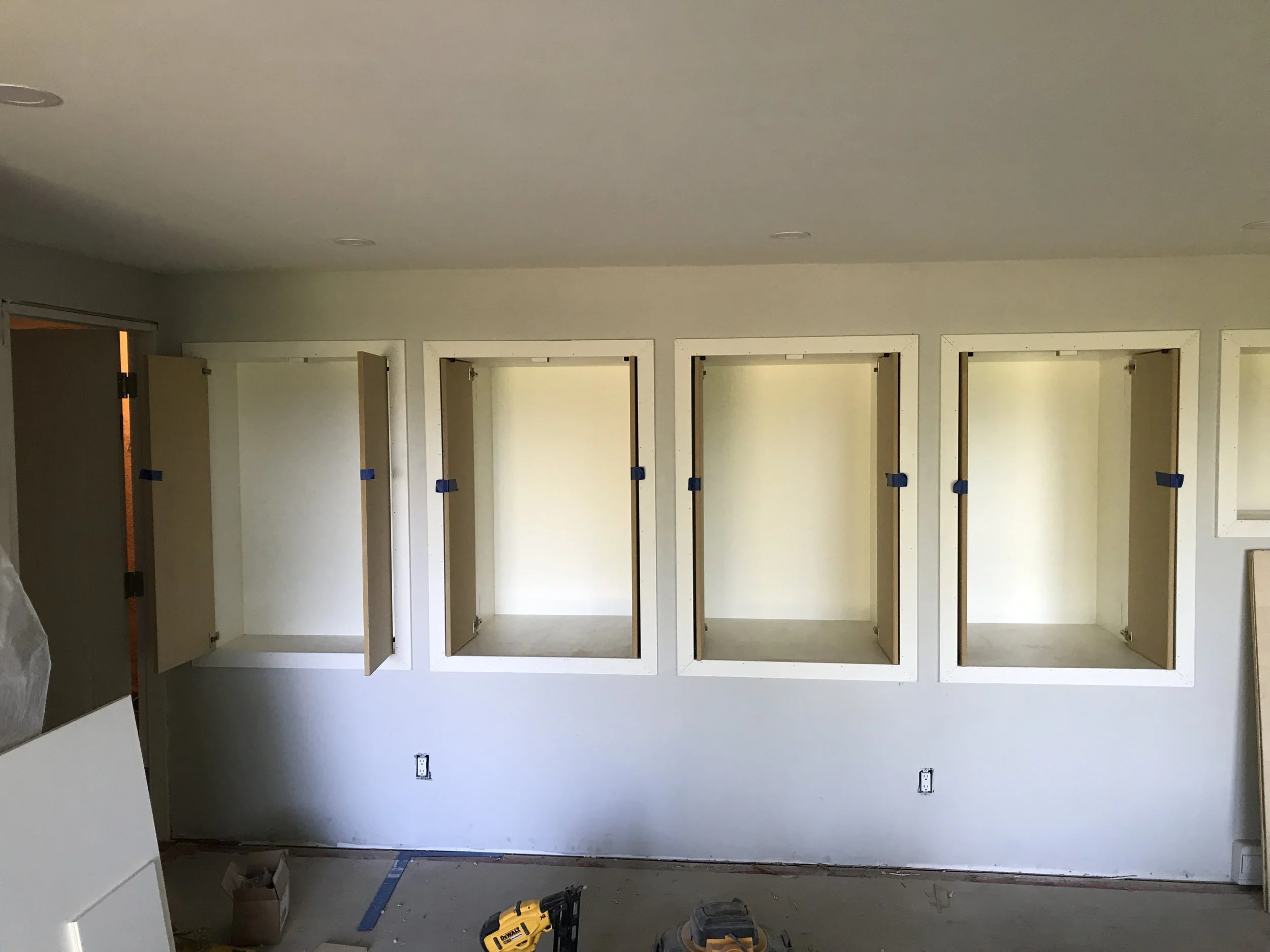 The room is under construction with framing for a wall installation. There are three door openings framed with white wood, and temporary blue and orange painter's tape is visible. The floor is unfinished, and tools such as a yellow power drill and a 