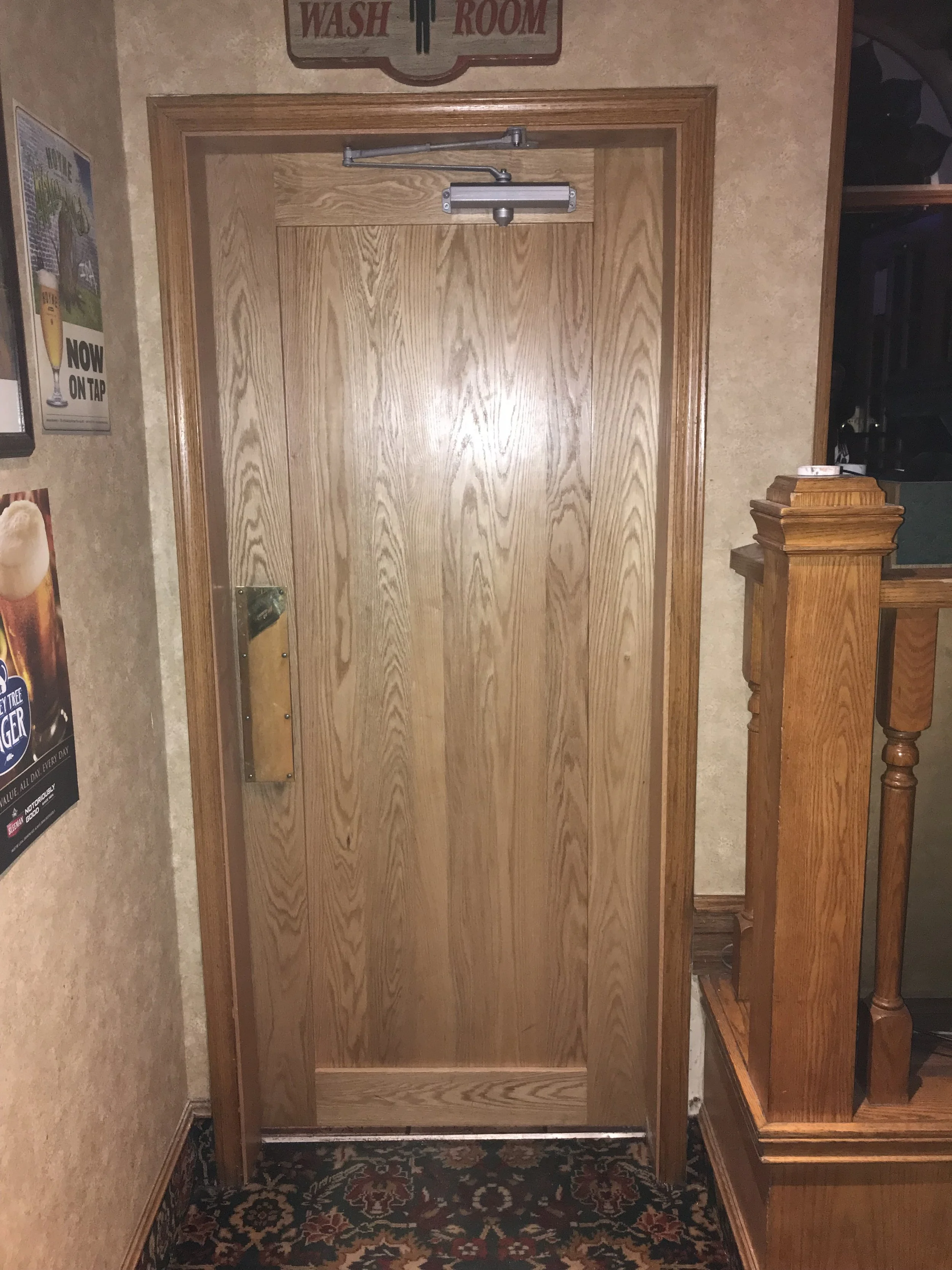 A wooden door with a push bar and a door closer at the top, set in a room with beige walls, a framed sign above the door that reads "WASH ROOM," and walls decorated with posters on the left. There is a patterned rug on the floor and wooden furniture 
