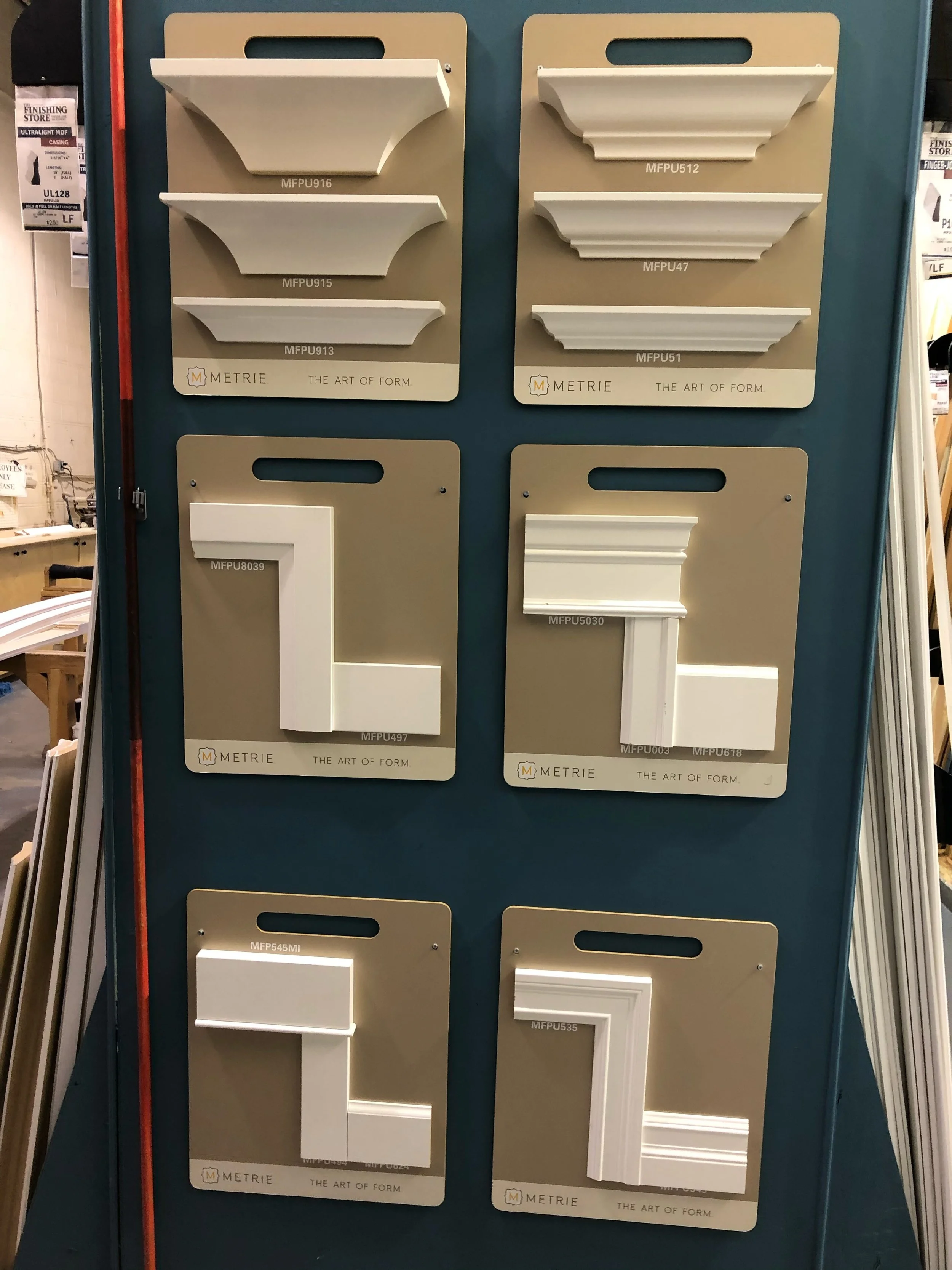 Display of decorative molding samples in various shapes and sizes on a retail board by Metrie, labeled with product codes.