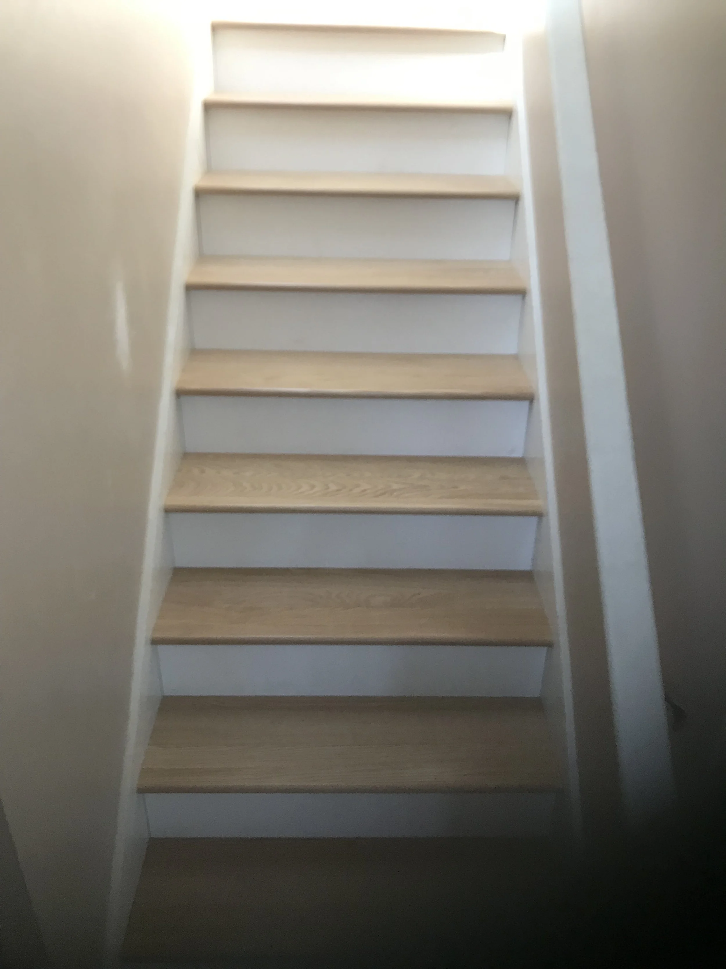 Wooden stairs with white risers viewed from bottom, with a bright light at the top illuminating the steps.