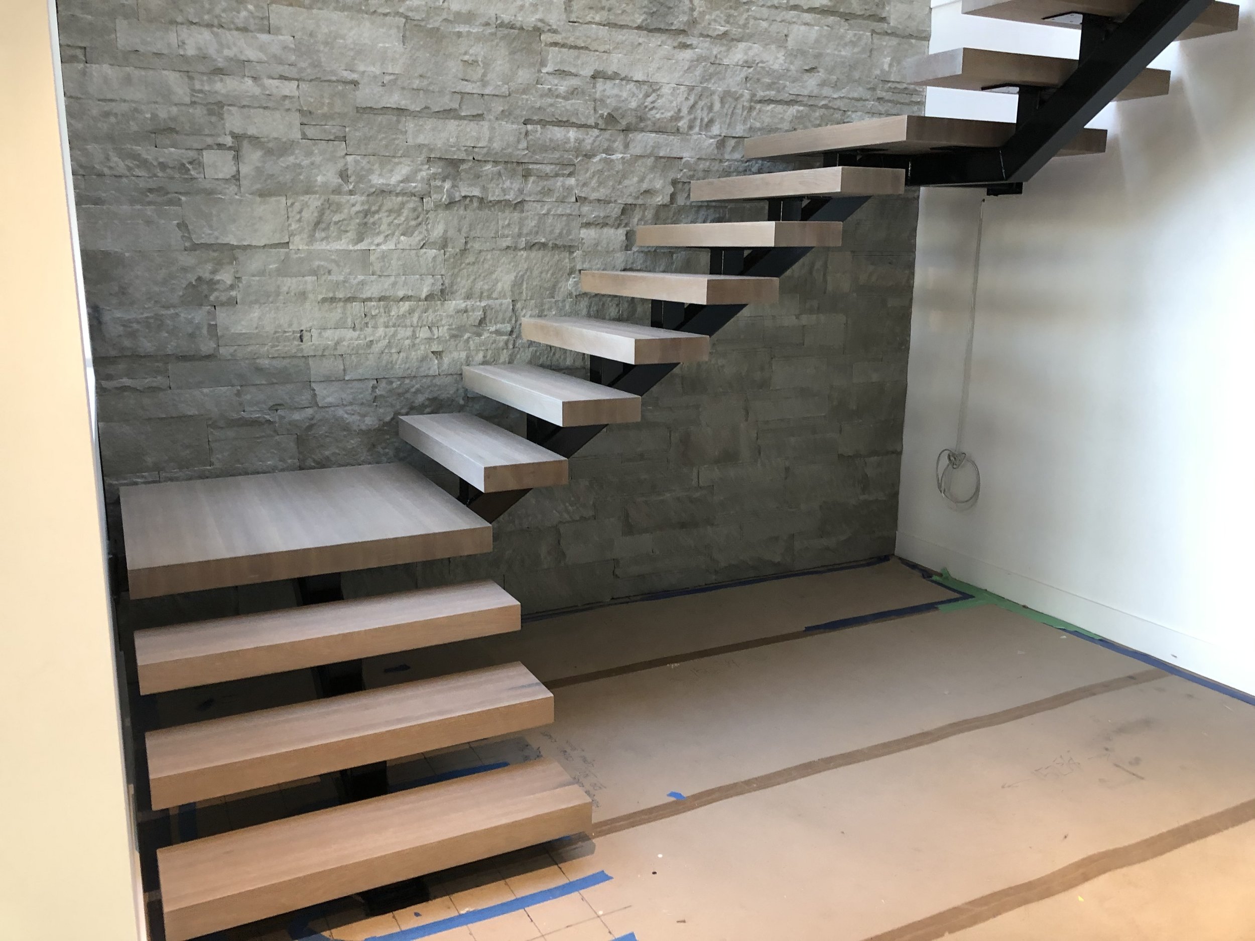 Interior of a building under construction featuring a floating wooden staircase with black supports against a textured stone wall.