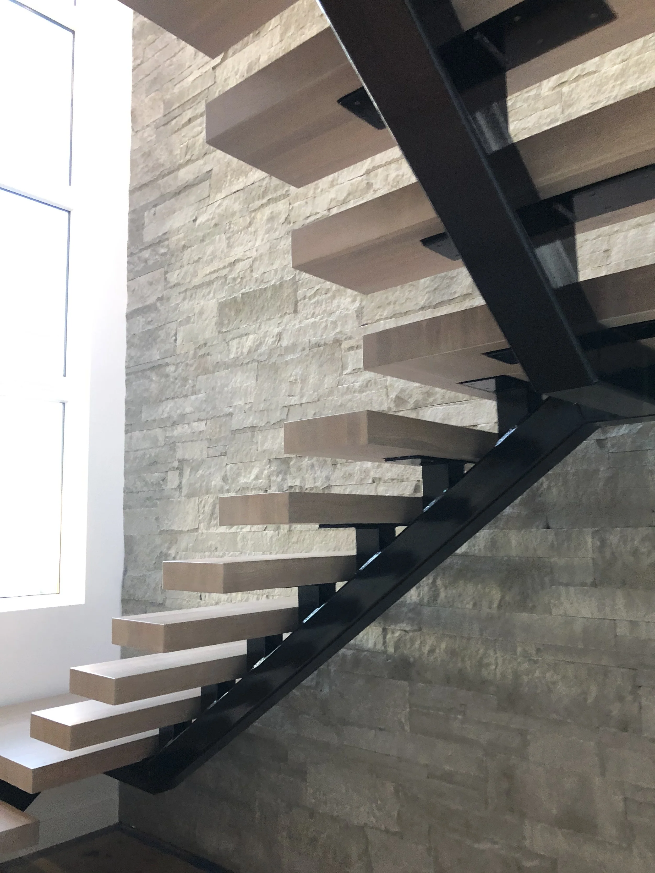 Interior staircase with wooden steps, black metal railing, stone wall on side, and a large window above.
