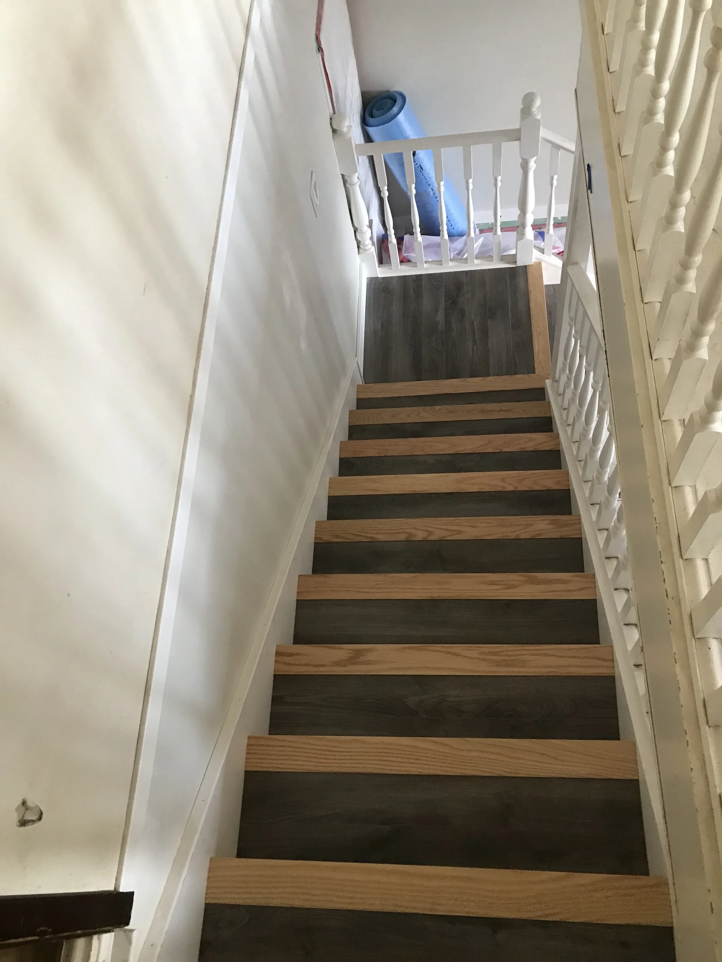 Top view of a staircase with alternating dark and light wooden steps and white railings, leading to a higher floor with a roll of blue material and some construction materials nearby.