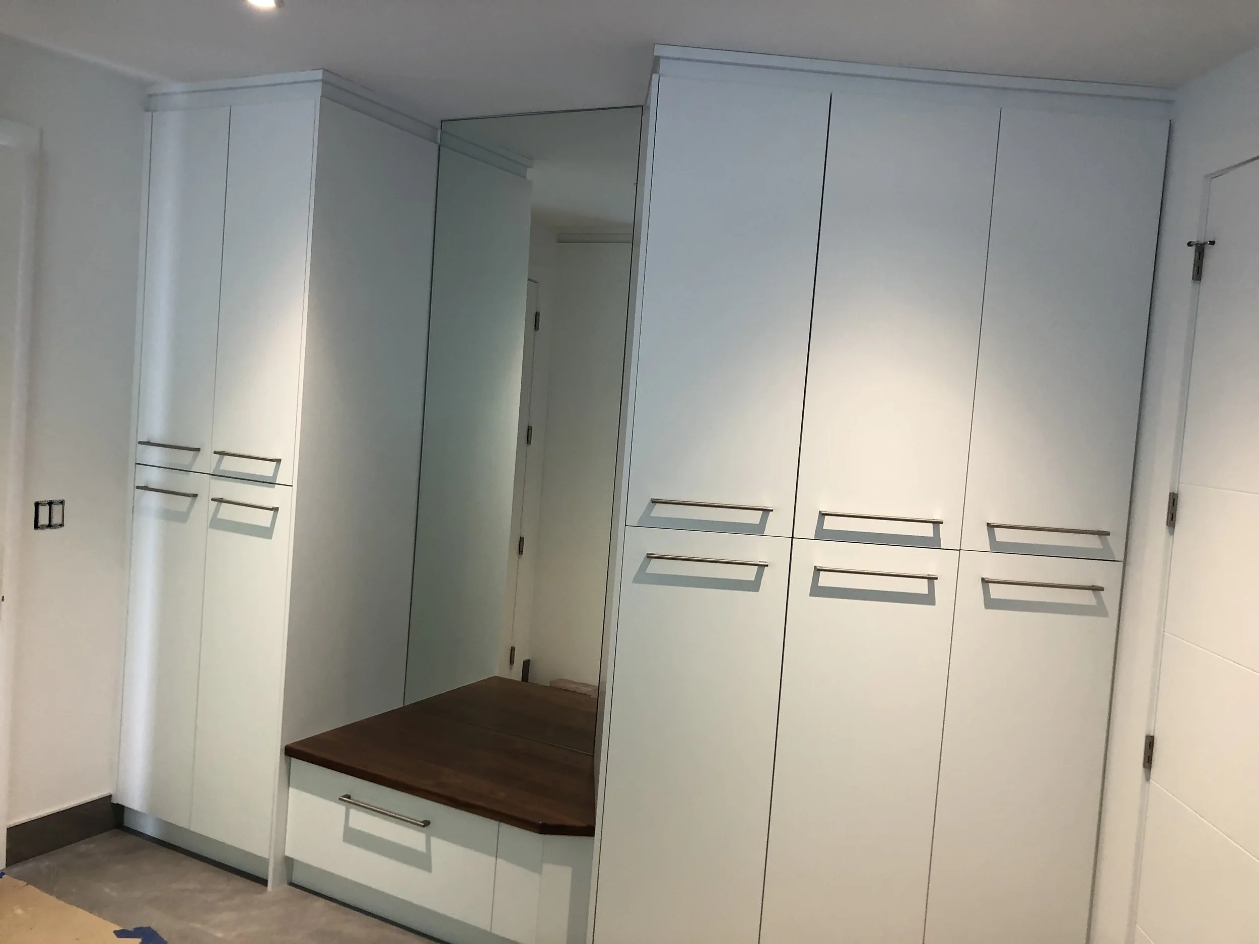 Kitchen cabinets with a wooden countertop and a mirror on one of the cabinet doors, white cabinet doors with silver handles, and a beige tiled floor.