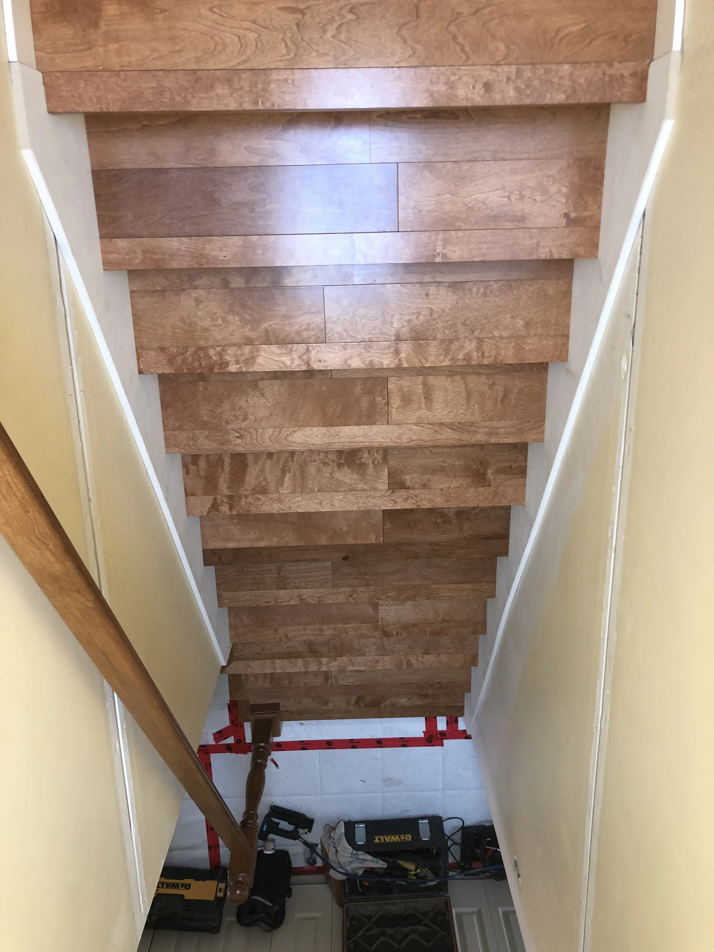 Indoor view of a staircase with wooden steps, walls painted yellow on both sides, white baseboards, and tools and equipment at the bottom, indicating ongoing construction or repair.