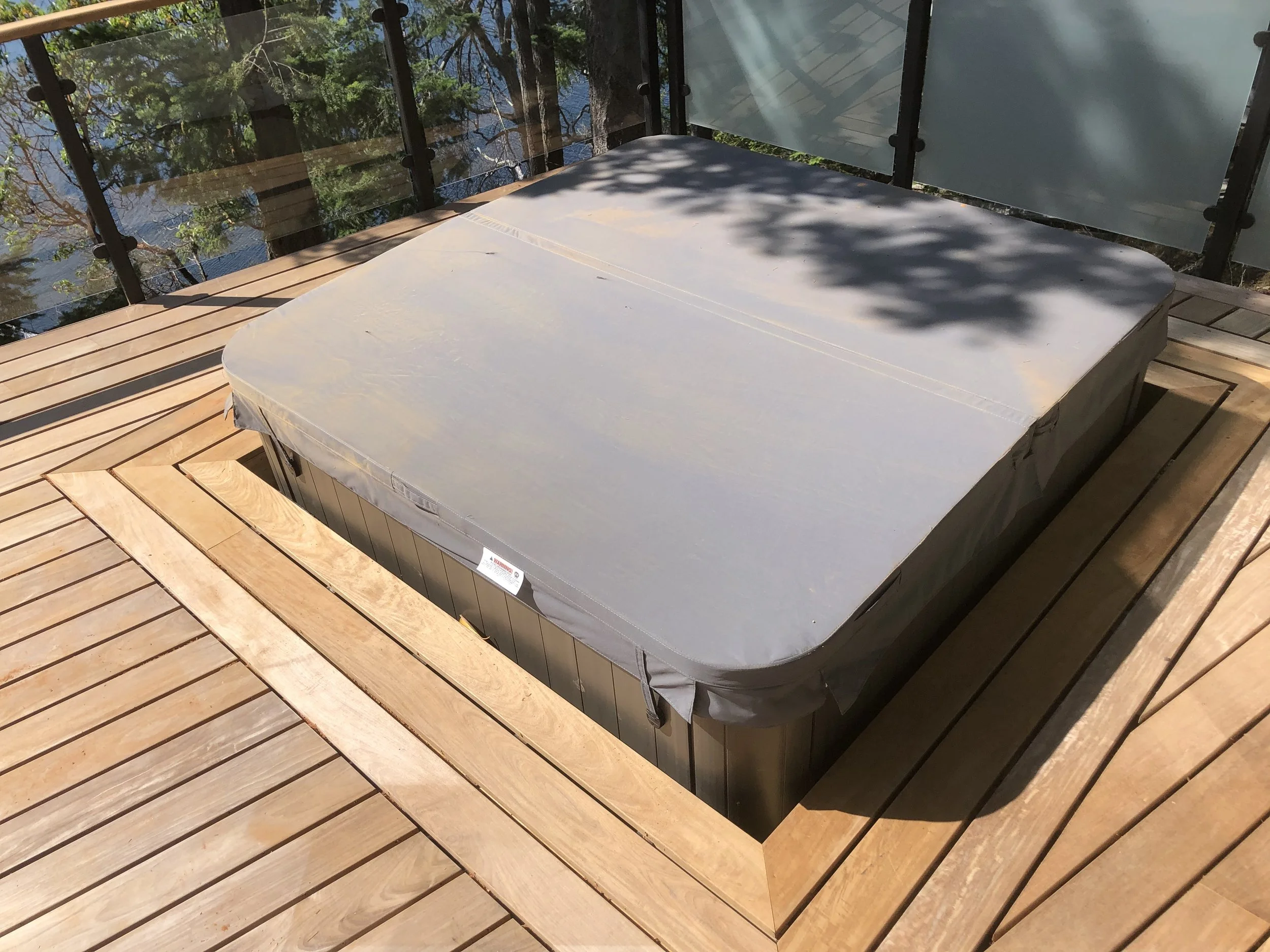 A hot tub covered with a gray cover on a wooden deck with a railing, surrounded by trees casting shadows.