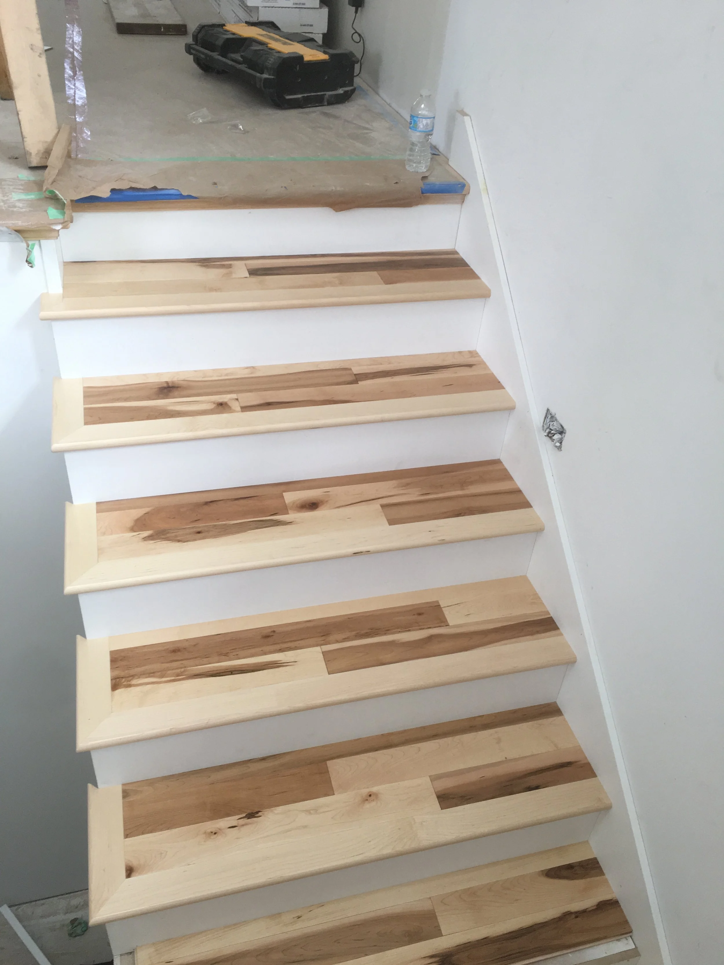 A set of wooden staircase steps is being built, showing progress from dark stained wood to light natural wood as the work continues. There is construction equipment and a water bottle visible in the background.