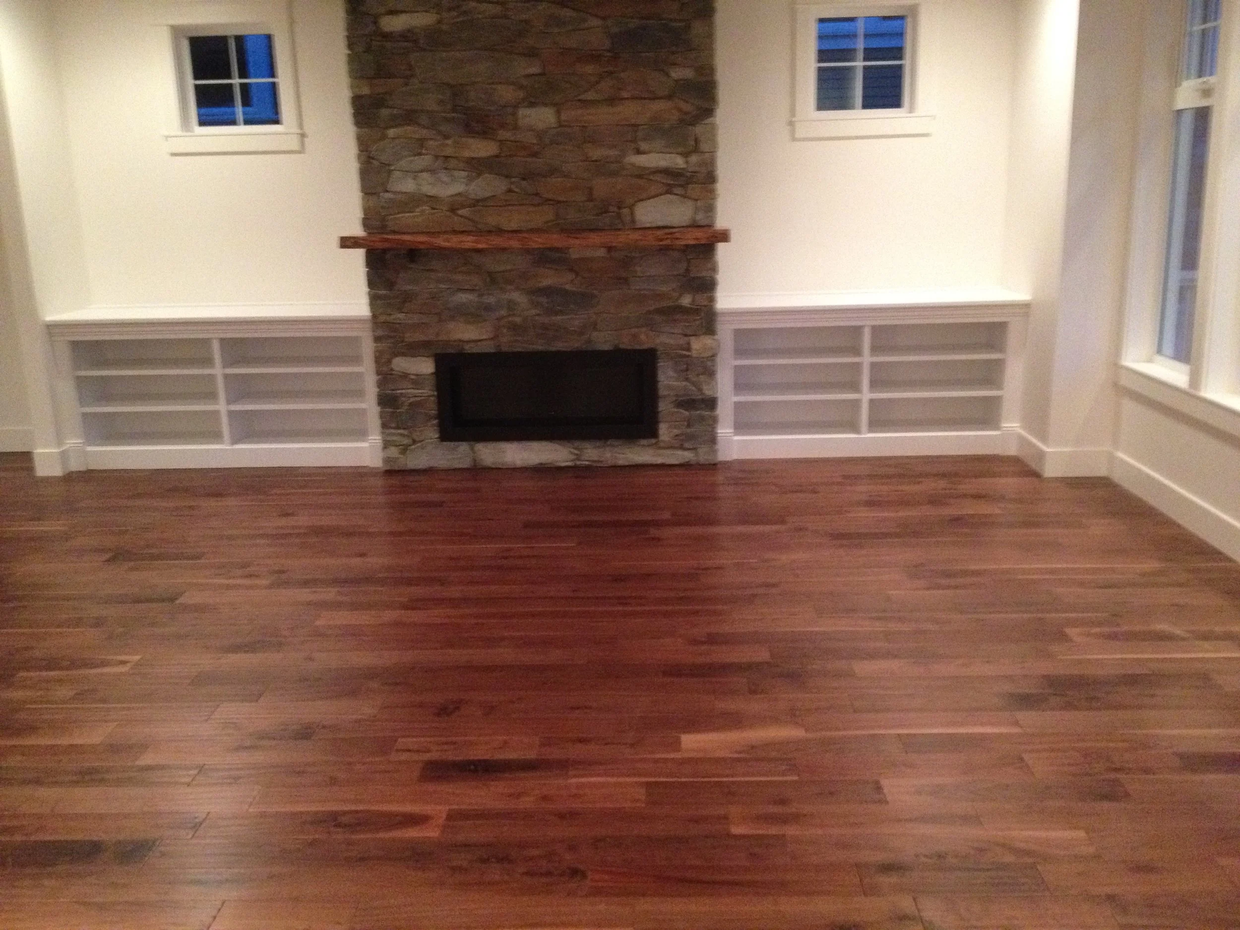 A room with wooden flooring, a central stone fireplace with a wooden mantel, and built-in white bookshelves on either side of the fireplace beneath two windows.
