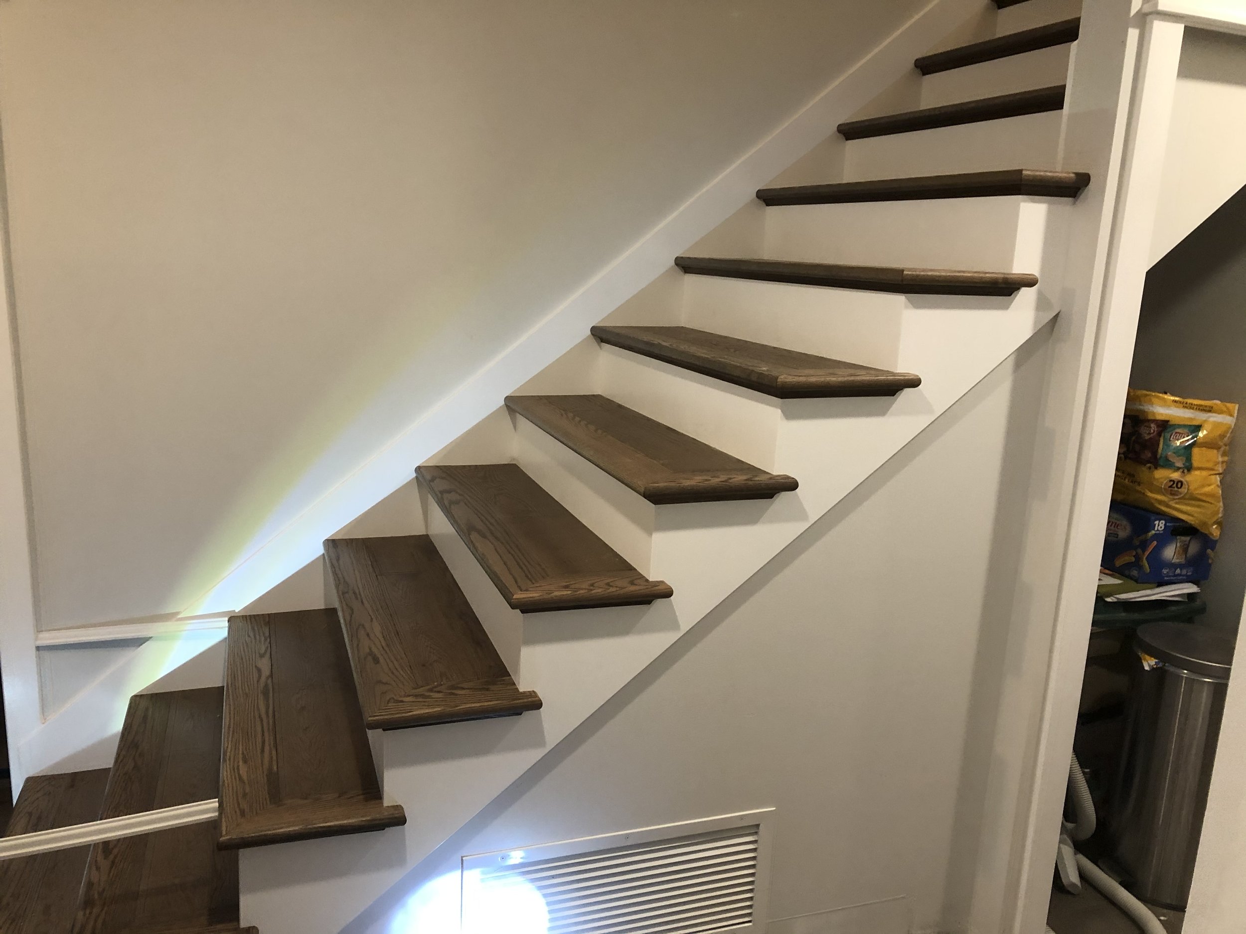 Interior staircase with dark wooden treads and white risers, ascending past a white wall. A vent is visible at the bottom of the wall, and some storage bags are seen on the right side of the image.