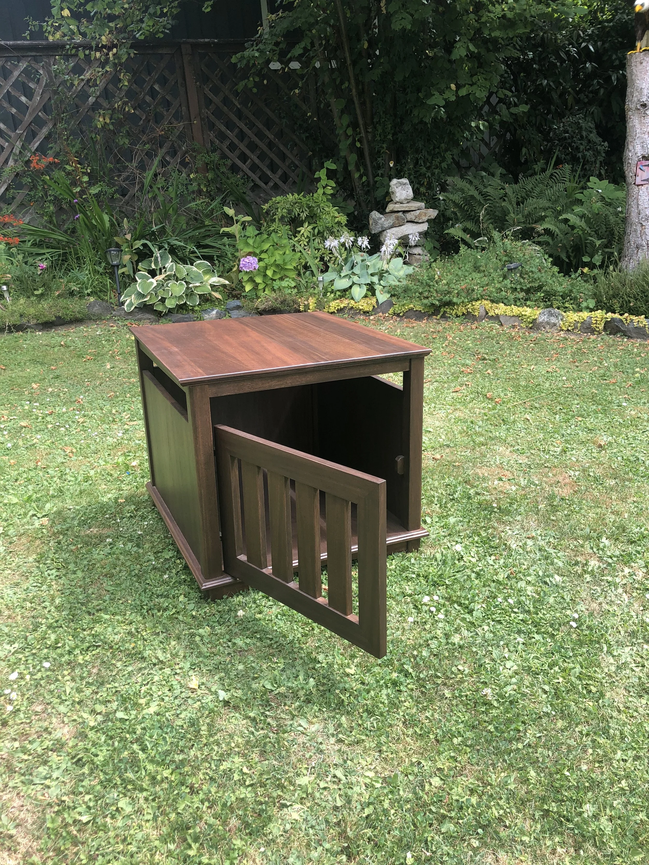 A wooden piece of furniture, possibly a table or cabinet, is placed on a grassy yard with a garden in the background. The garden includes various plants, flowers, and a small stack of rocks.