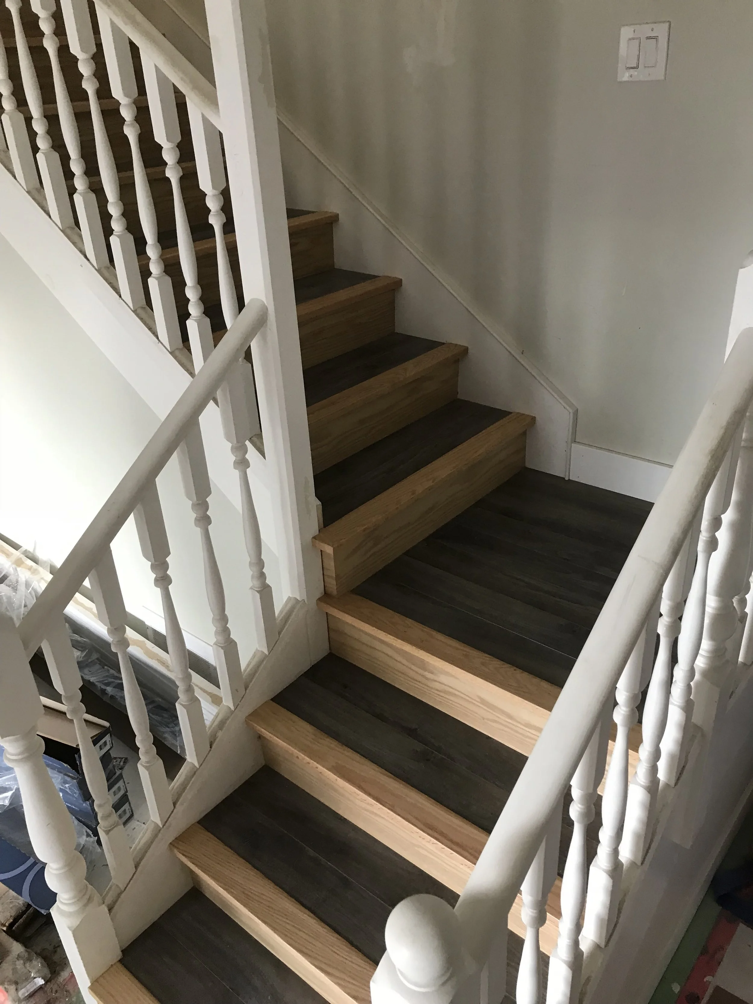 Interior view of a staircase with wooden steps painted in dark and light wood tones, white spindles and handrails, and a wall with a light switch, under construction or renovation.