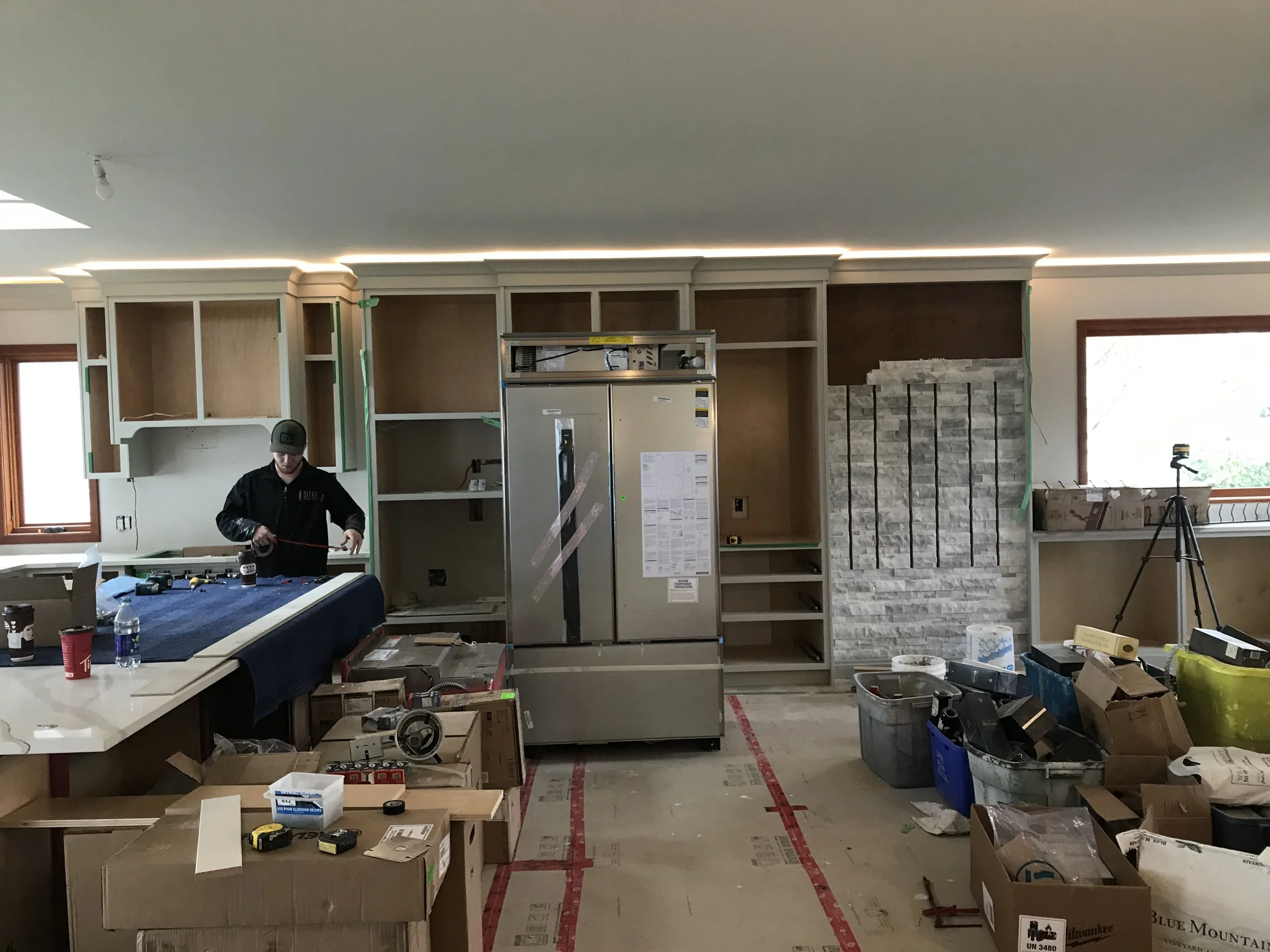 A person working on a kitchen renovation or installation, surrounded by various tools, materials, and boxes, with partially built cabinets and a stone accent wall in the background.