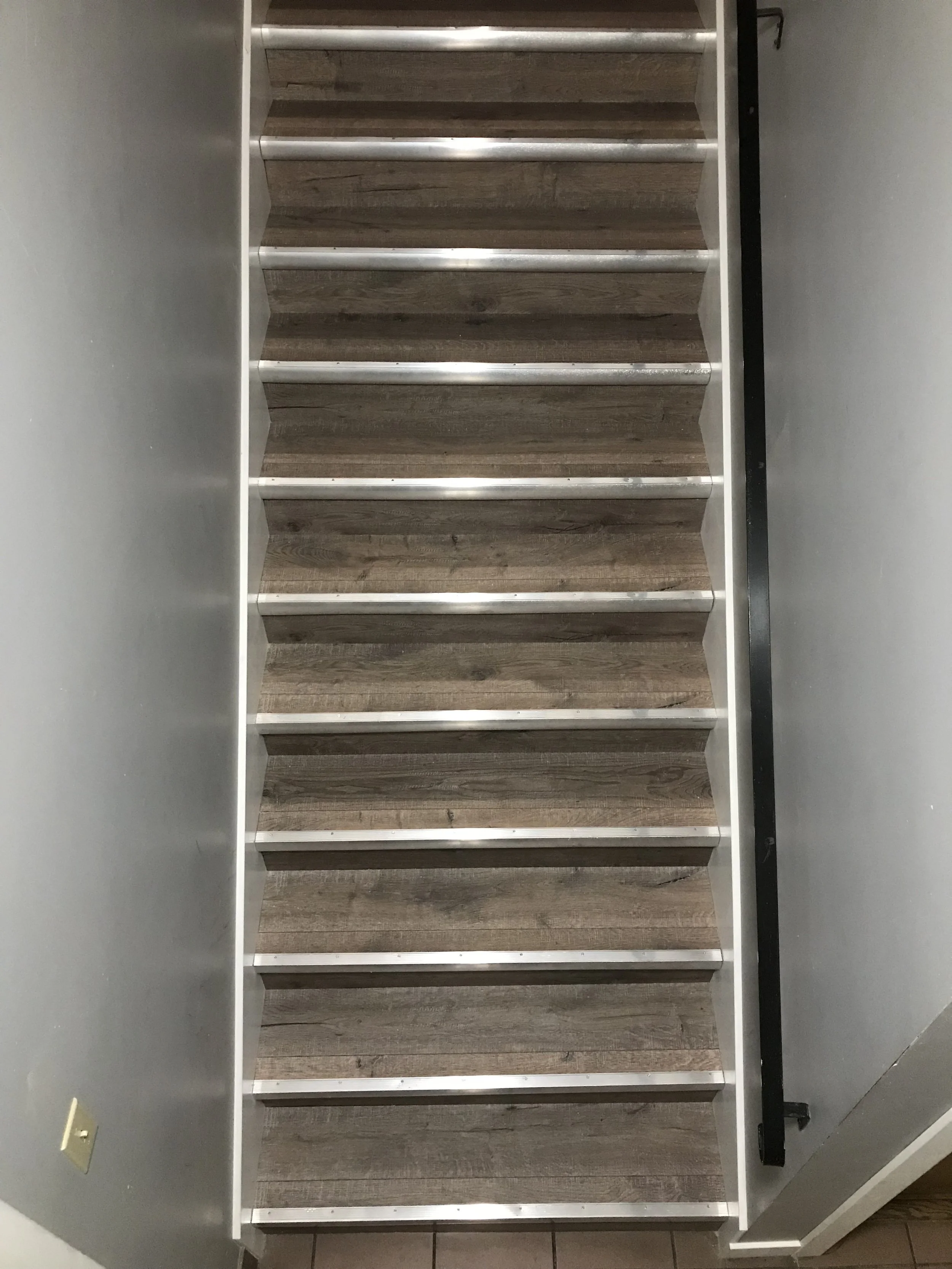 Indoor staircase with wood-look steps, white risers, and a black handrail along the right side. The wall on the left side is painted gray, and the floor at the base of the stairs has brown tiles.