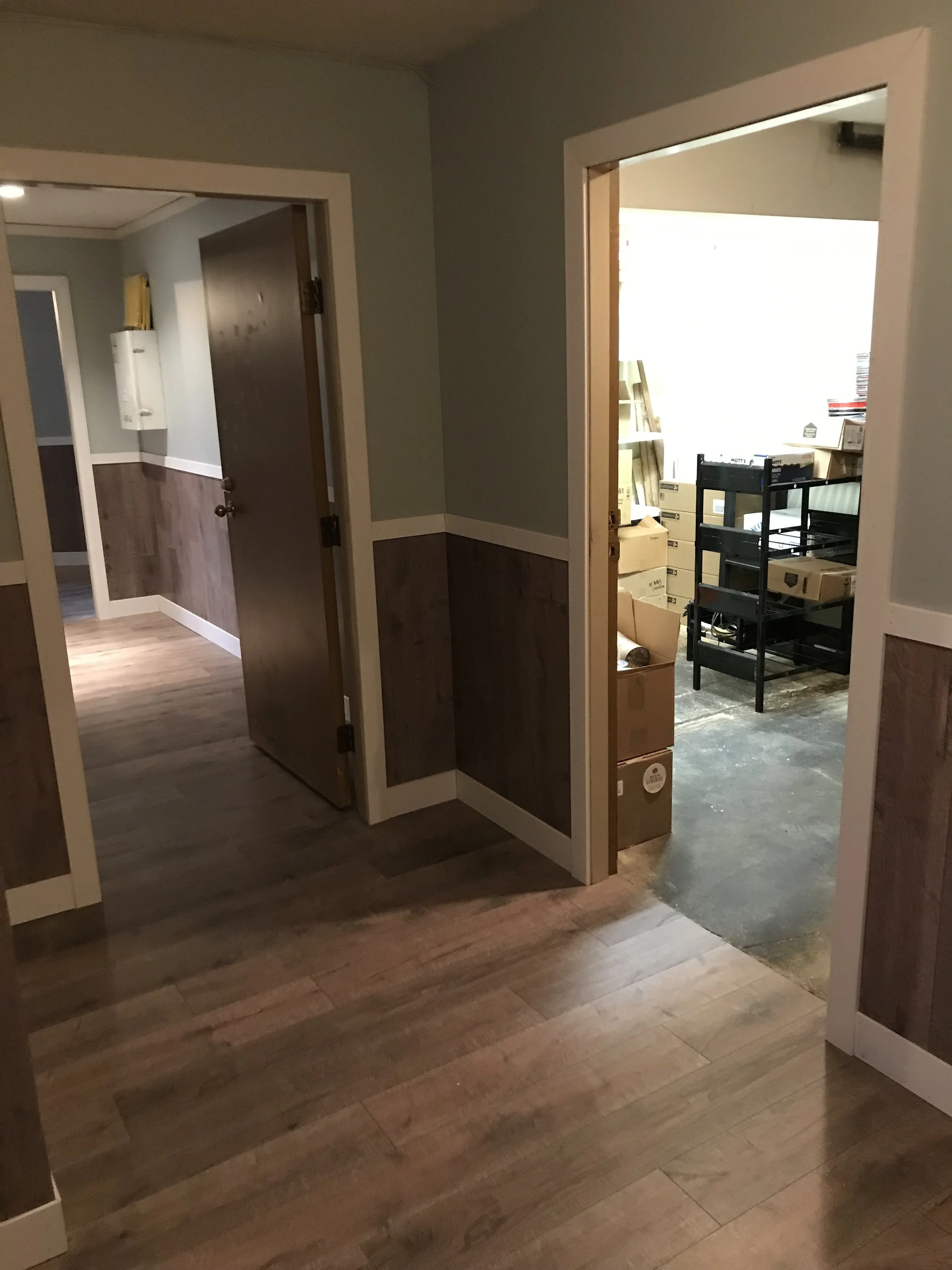Interior space with wooden flooring, multiple doorways leading to other rooms, and a storage or utility area with boxes and shelves visible through one doorway.