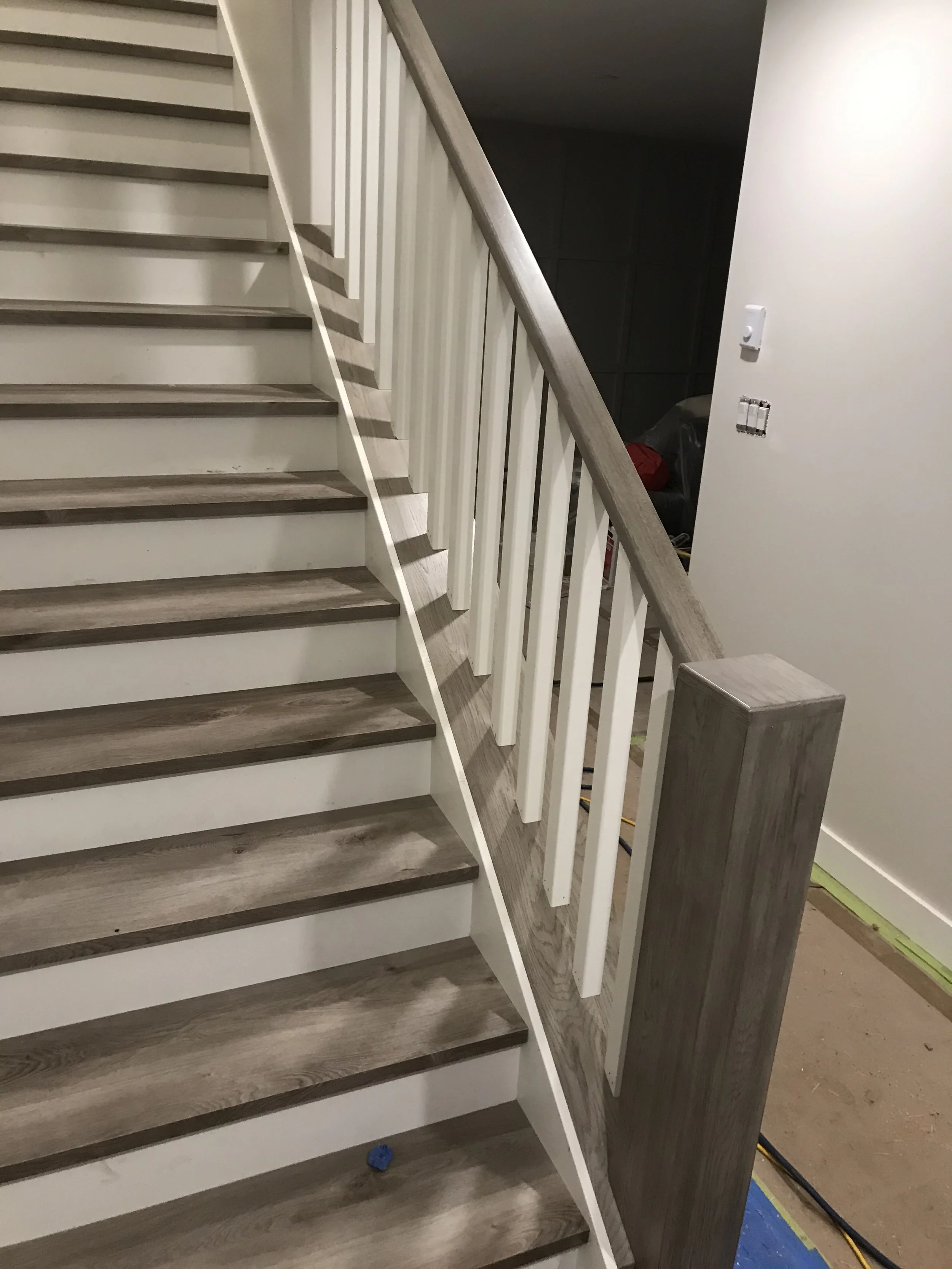 A staircase with wooden steps and white risers, white balusters, and a wooden handrail. Partially visible construction area with tools and materials.
