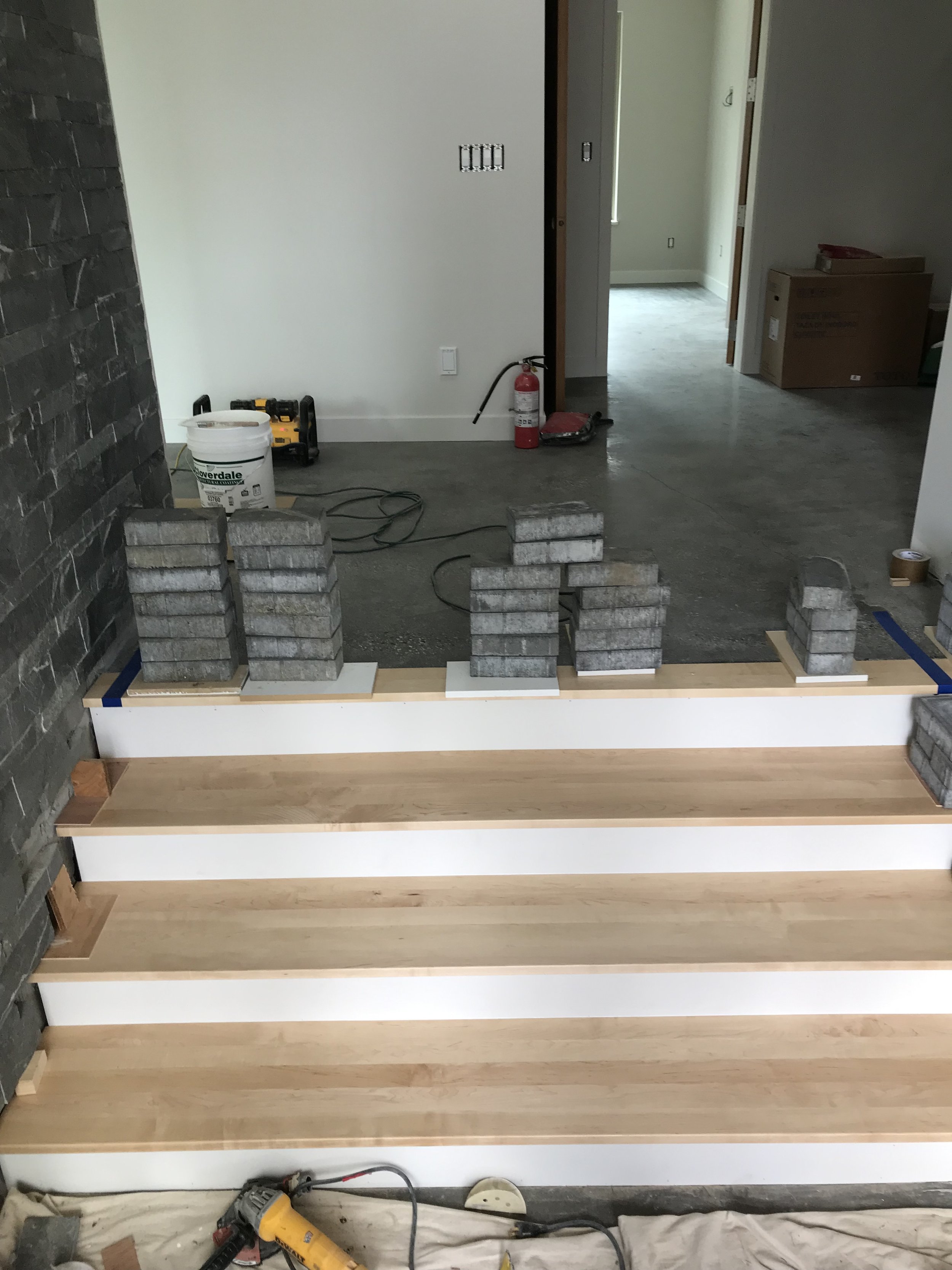 Interior room of a house under renovation with wooden stairs, construction tools, and building materials visible. The space appears to be in the process of flooring installation.