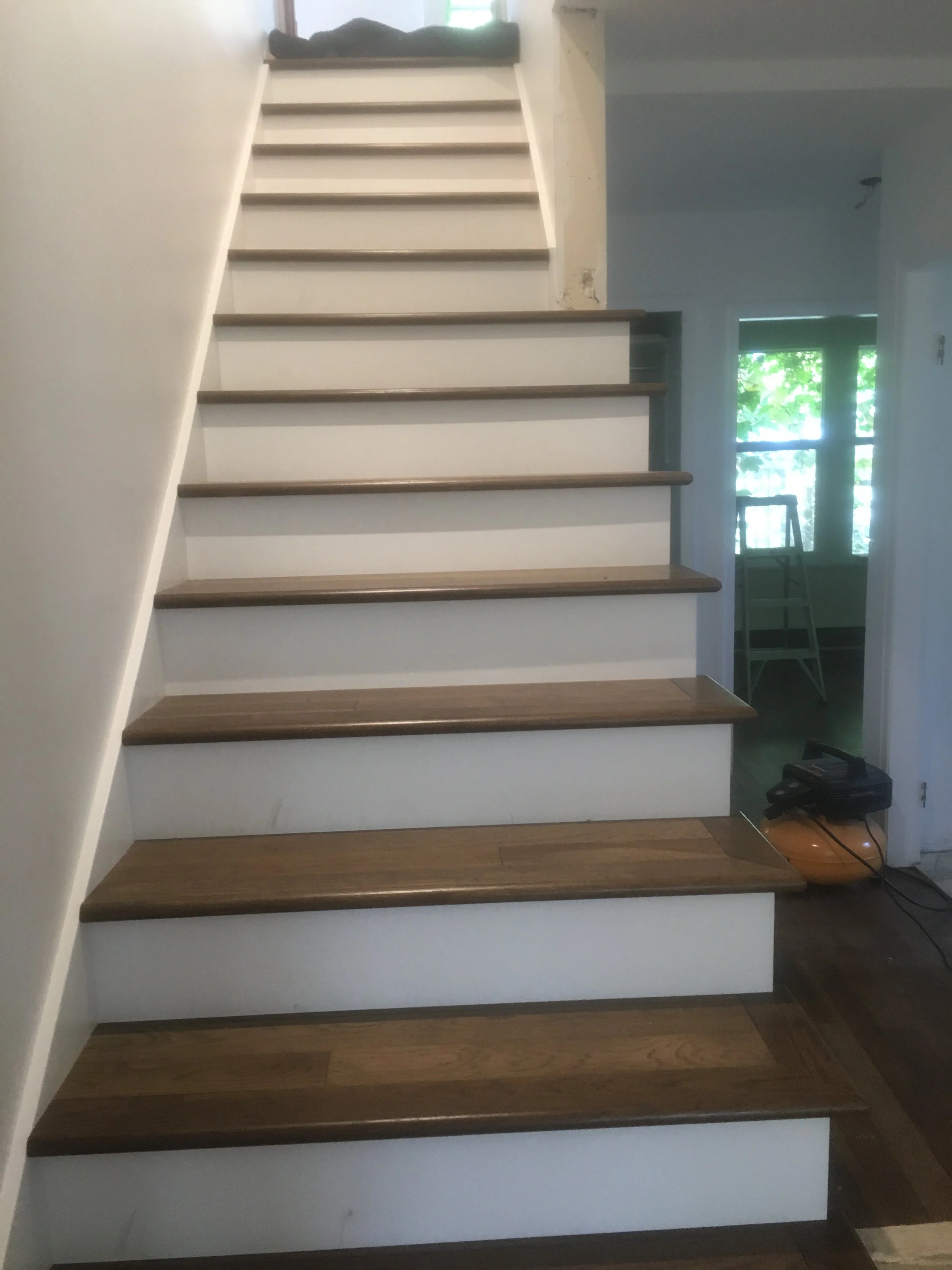 A staircase with wooden steps and white risers inside a home. There is a window at the top of the stairs with some greenery visible outside, and a ladder is visible through an open doorway on the right side of the image.
