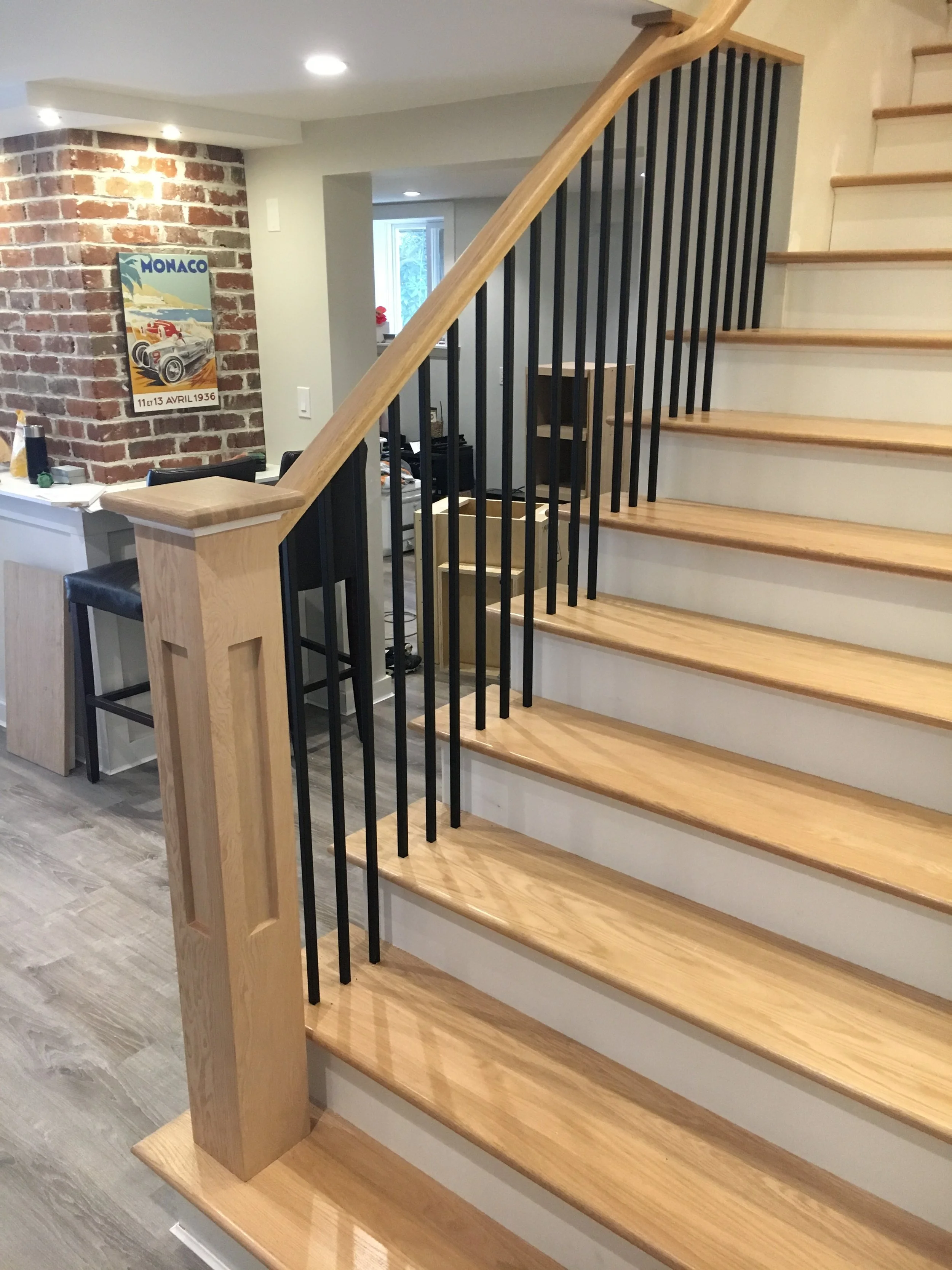 Interior view of a staircase with wooden steps and black metal balusters, adjacent to a brick wall with a vintage Monaco racing poster, in a home with recessed lighting.